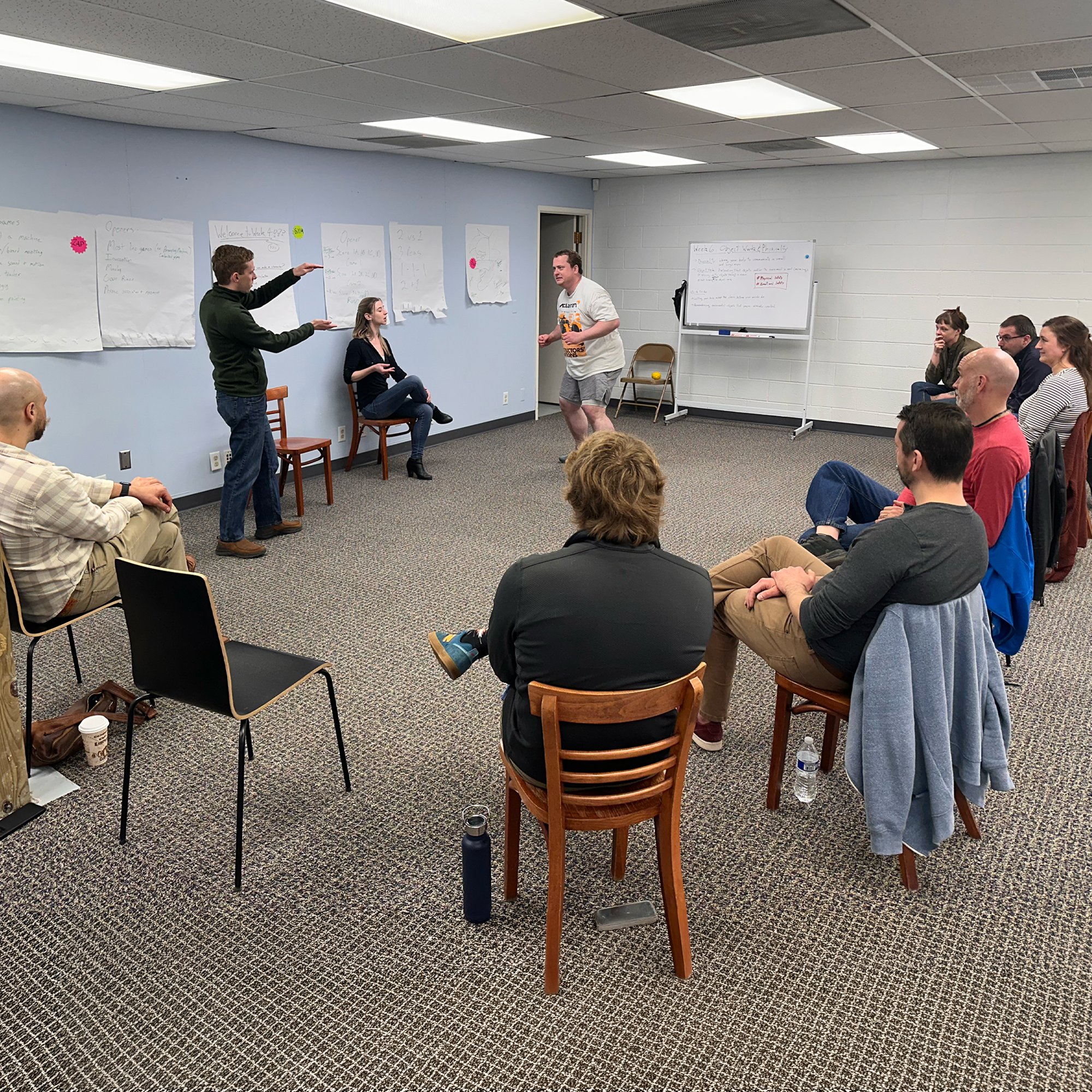 A group of people participating in a workshop or meeting in a room with white walls, sitting on chairs arranged in a semi-circle. Two presenters are at the front, one standing and one sitting, engaging with the audience. There are large sheets of paper with notes and diagrams on the walls, and a whiteboard in the background.
