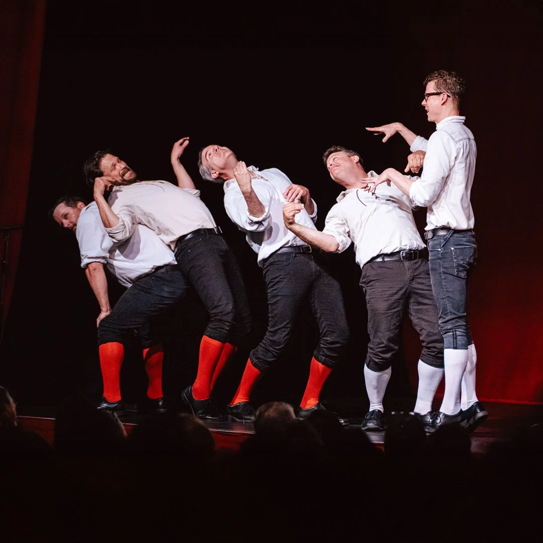 Group of five men performing a humorous or theatrical act on stage, with exaggerated poses and expressions, wearing white shirts, dark pants, and colorful socks.