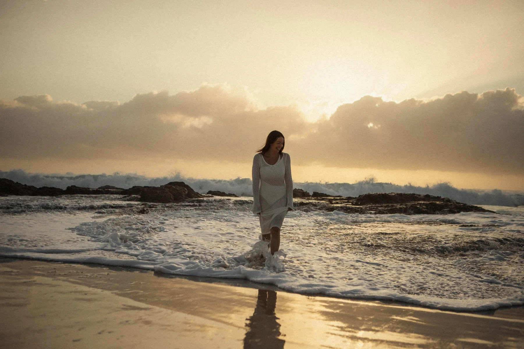 A woman in a white dress walking through ocean waves during sunset, with rocky formations in the background and the sky partly cloudy.
