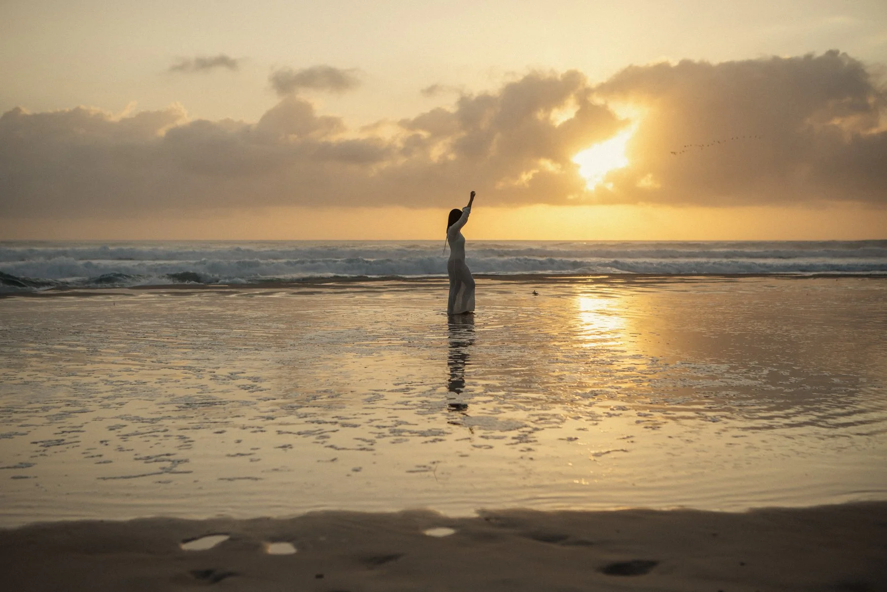 A woman in a white dress standing in shallow water on a beach at sunset, with her arm raised and clouds partly covering the sun.
