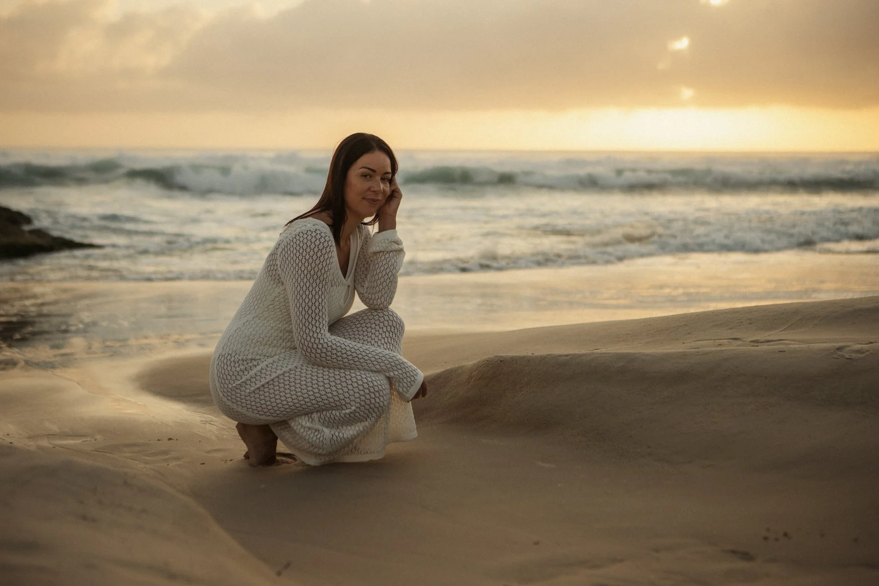 Woman in white crochet dress kneeling on sandy beach at sunset with ocean waves in the background.