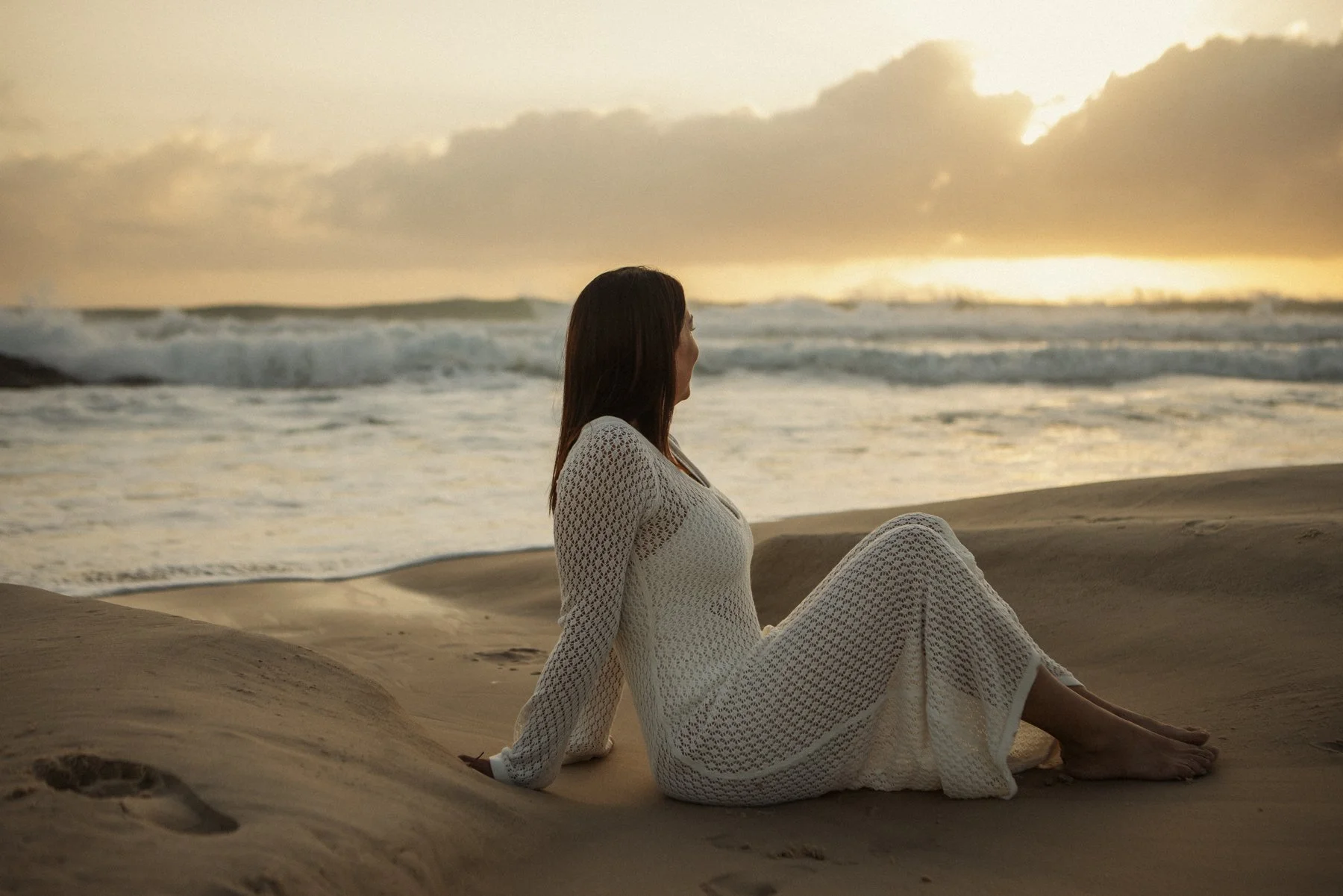 A woman in a white dress sitting on the sand at the beach during sunset, facing the ocean.