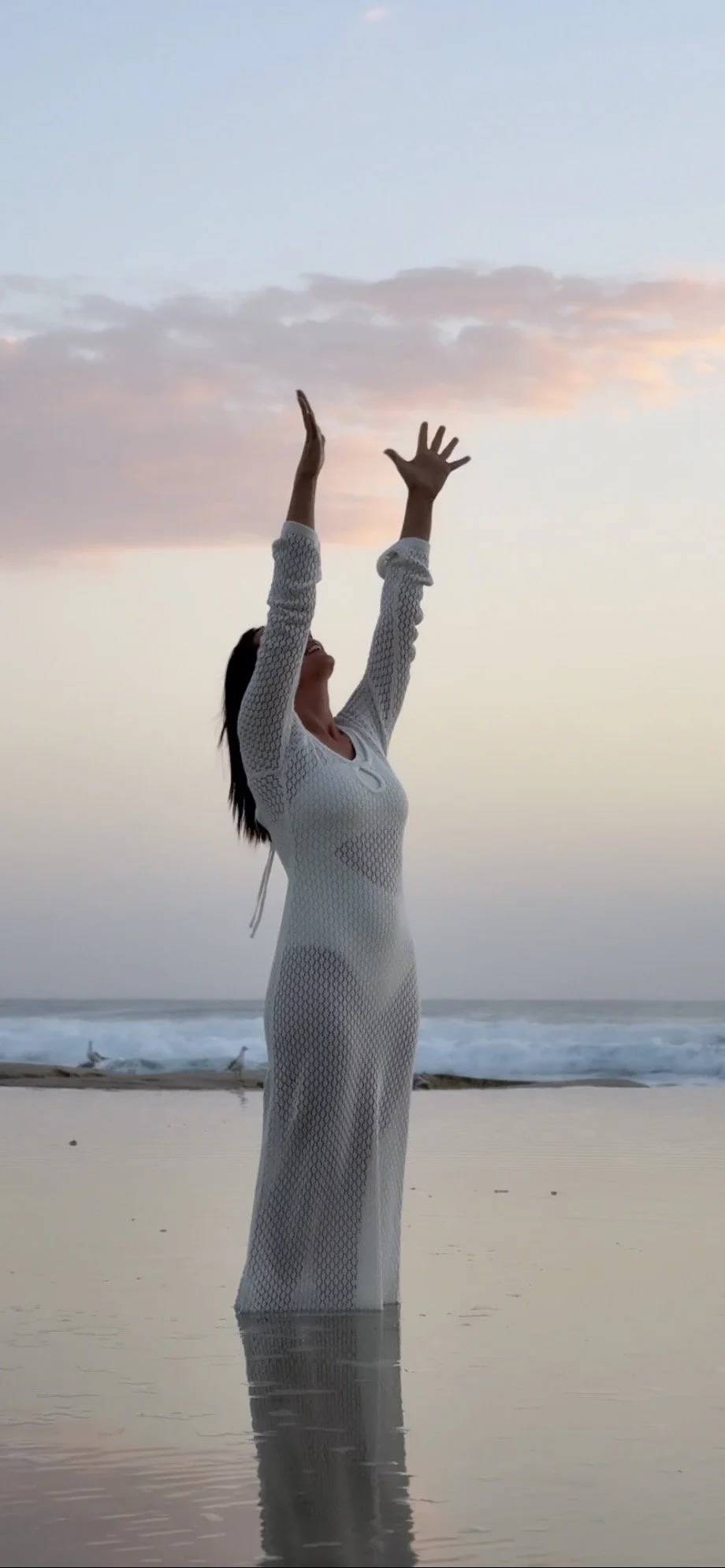 A woman in a white, long-sleeved, semi-transparent dress standing in shallow water on a beach at sunset, with her arms raised upward and the sky with pink and blue hues in the background.