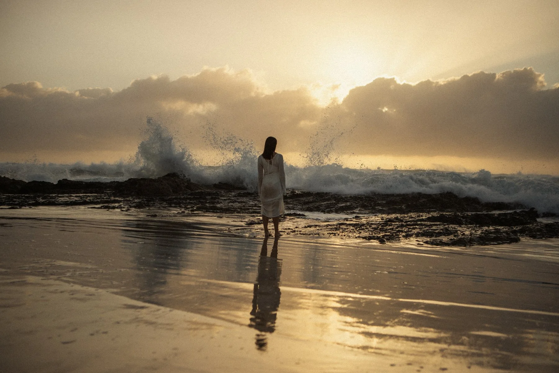 A woman in a white dress standing on the beach near the water with waves crashing behind her during sunset, with her reflection visible on the wet sand.