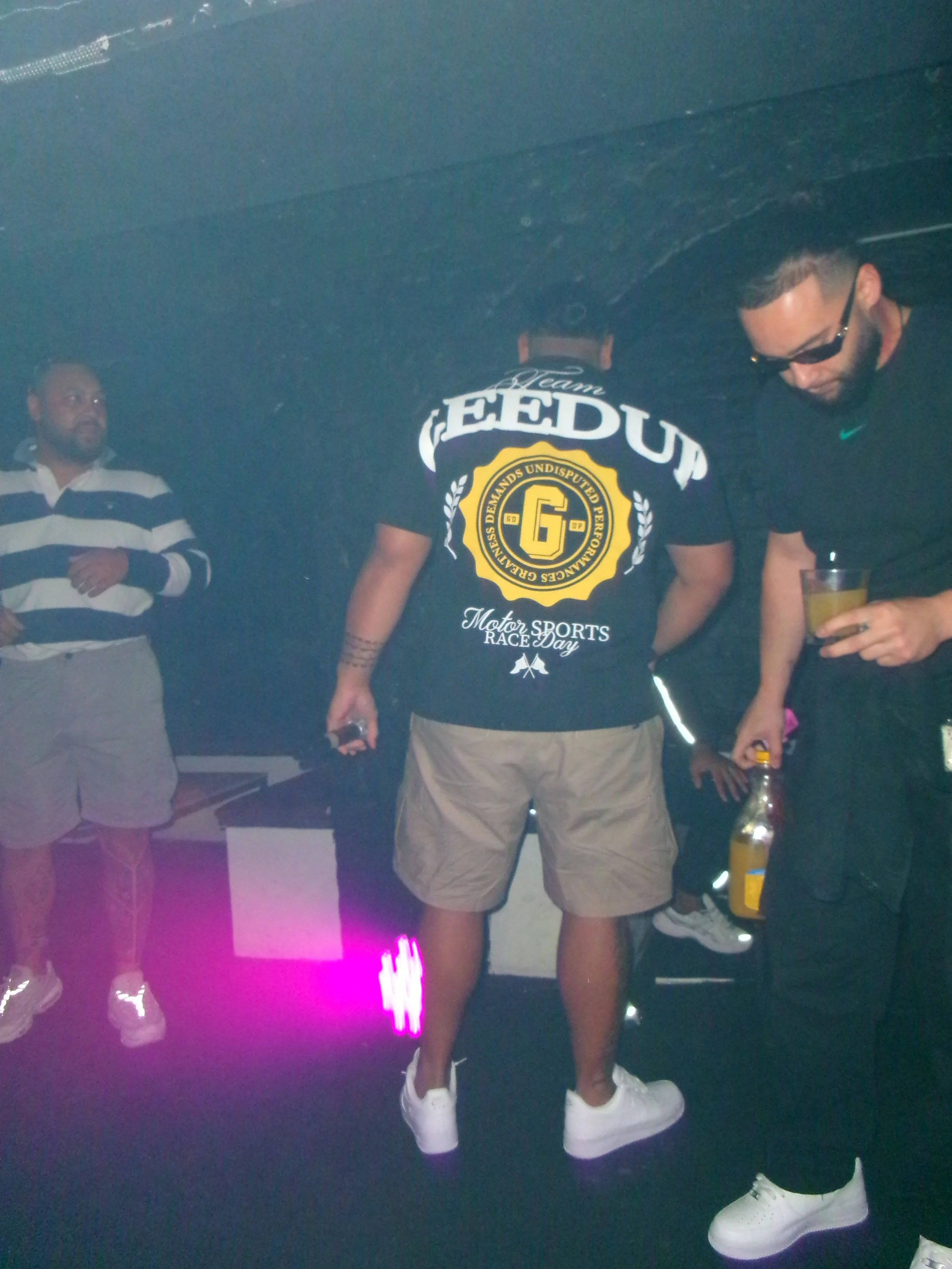 Three men at a party or nightclub. One man in the foreground wears sunglasses, a black shirt, and holds a drink. Another man in the background wears a shirt with 'FEED UP' on the back. The third man is on the left in a striped shirt with hands folded.