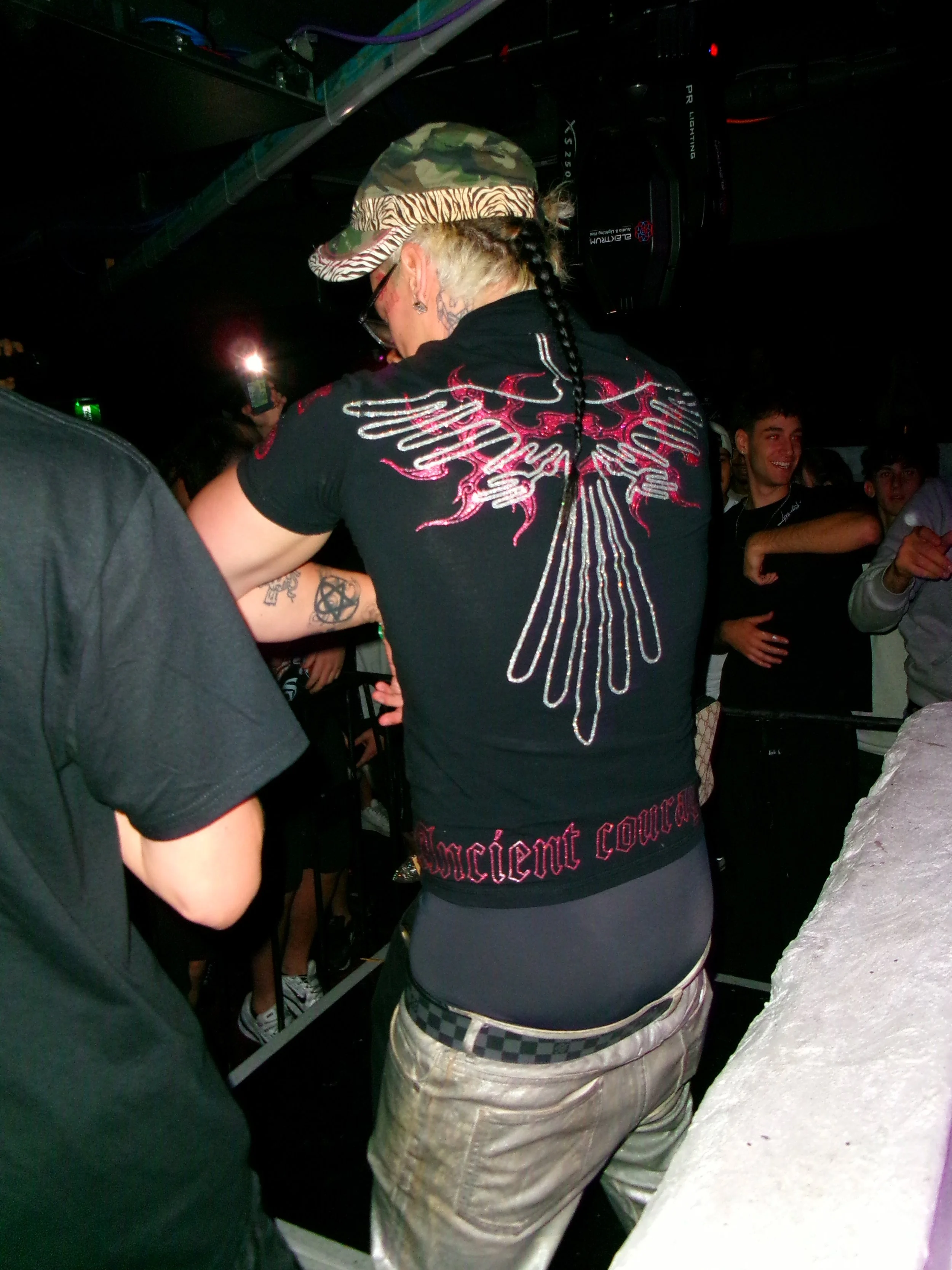 Person with platinum blonde hair, wearing a camouflage hat, black shirt with red and white graphics on the back, and beige pants, standing among a crowd at a dance or concert.