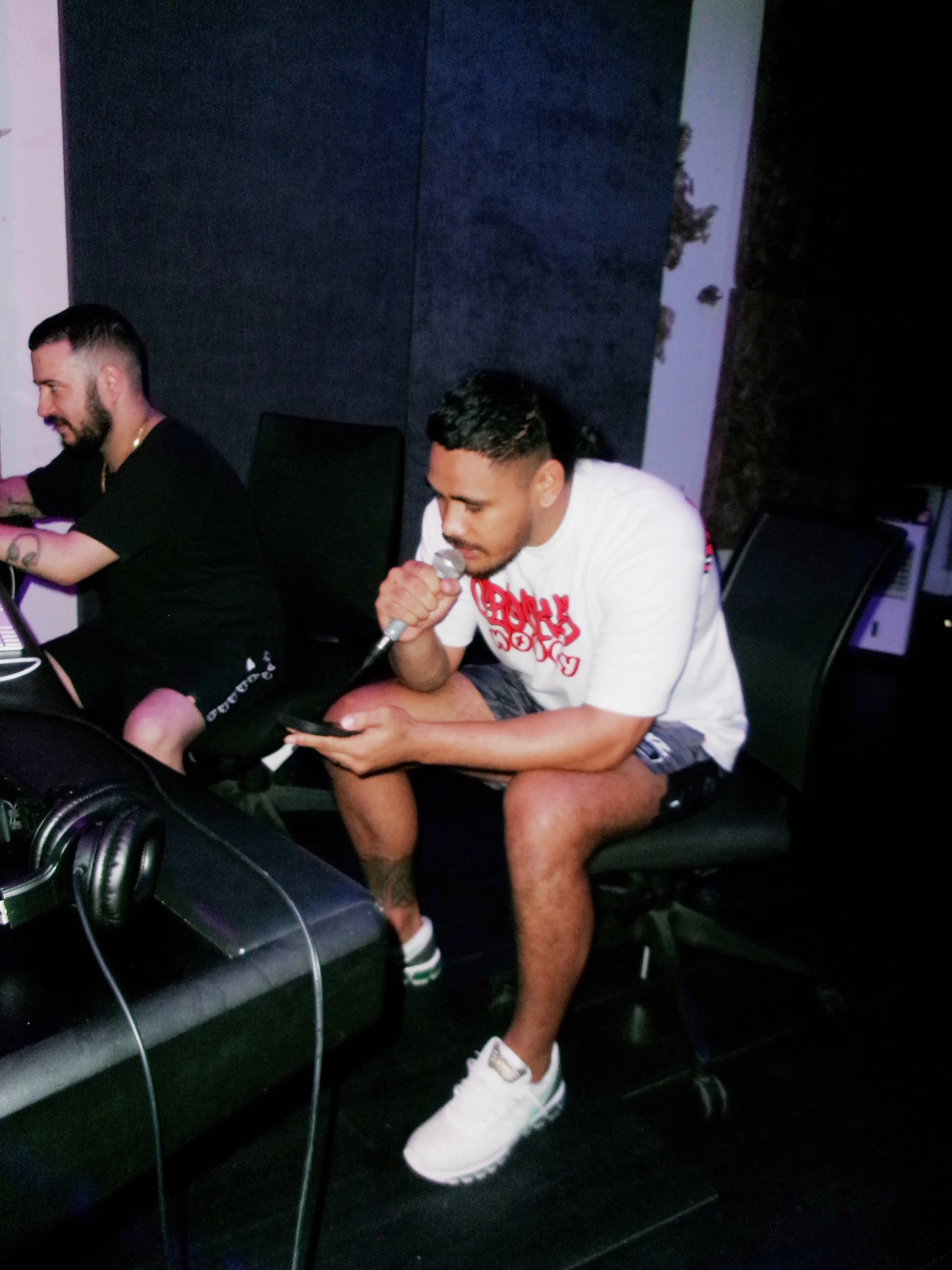 A man sitting on a chair in a dimly lit room, holding a microphone in his right hand and a phone in his left, wearing a white t-shirt, black shorts, and white sneakers, with two tattoos visible on his left leg, and two other people nearby.