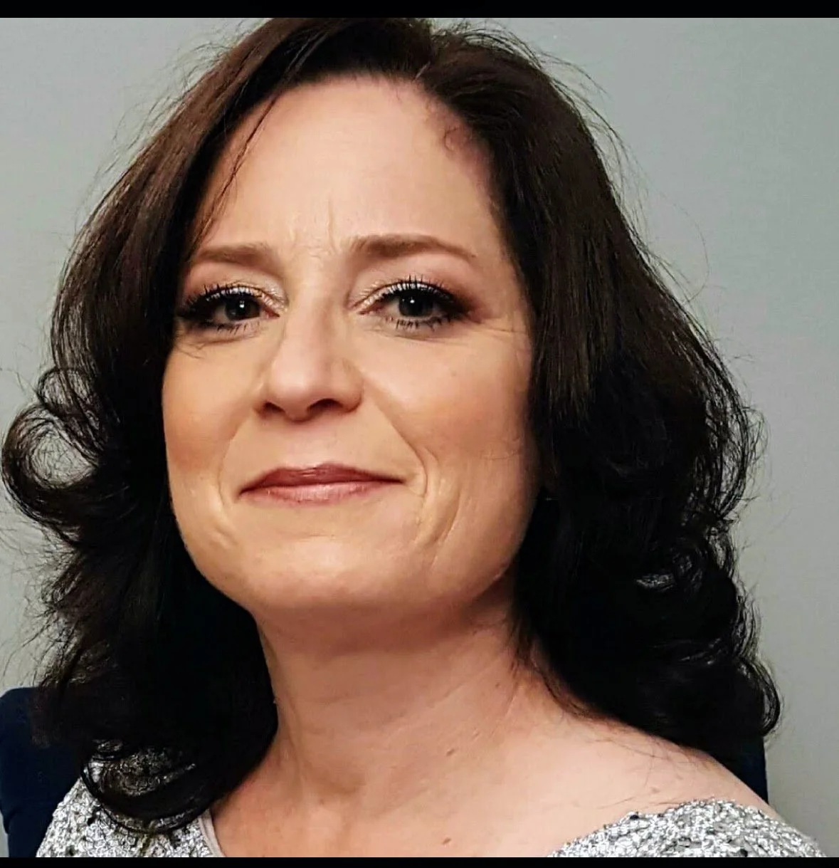 Close-up photo of a woman with dark, wavy hair, wearing makeup, and a silver top, smiling softly against a plain background.