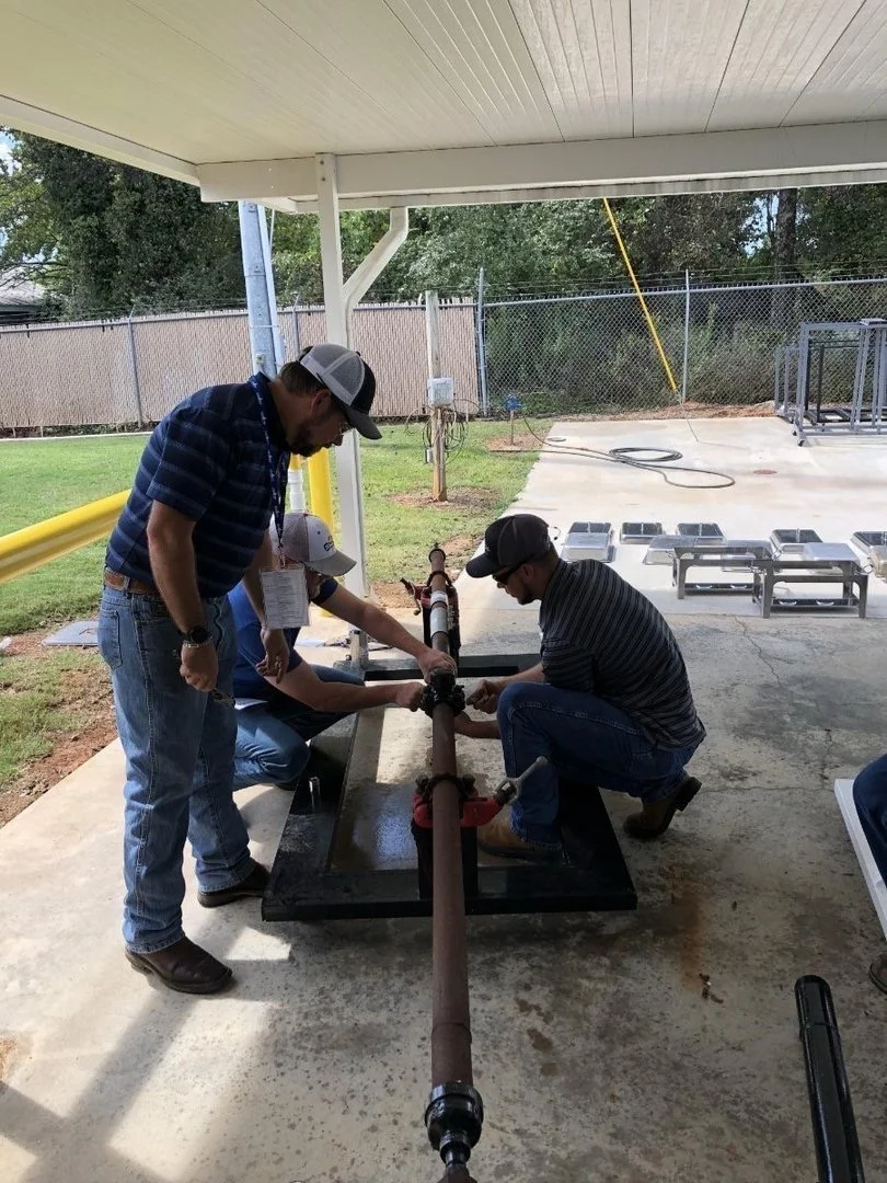 Three men working together on a large metal pipe outdoors, under a covered area with lawn and chain-link fence in the background.