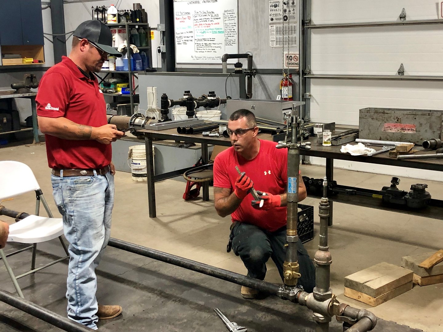 Two men working on plumbing pipes in an industrial workshop. One man is standing, holding a pipe, while the other is kneeling, working with the pipe fittings. Tools and materials are scattered around a workbench and shelves in the background.