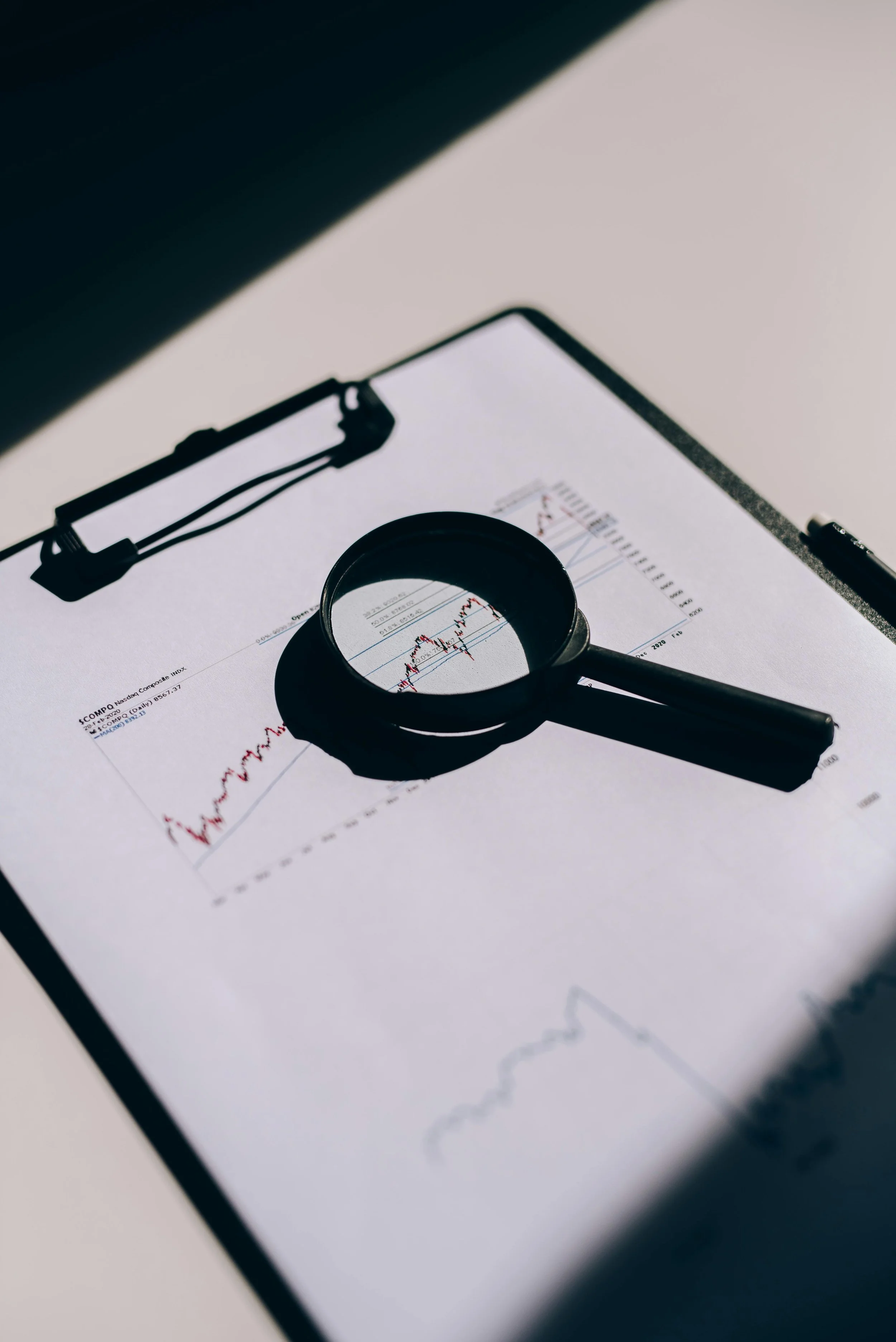 A magnifying glass placed over a financial chart showing stock or market data on a clipboard.