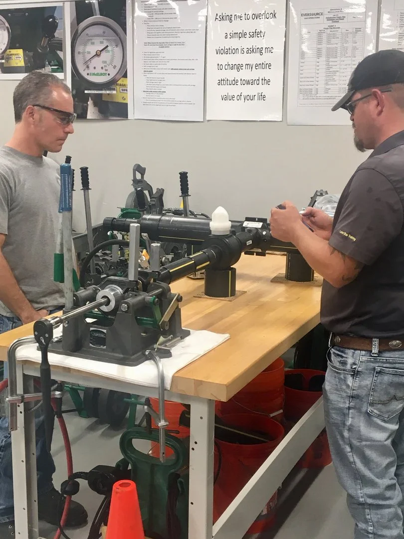 Two men working with a pipe threading machine at a workshop, with safety posters on the wall behind them.