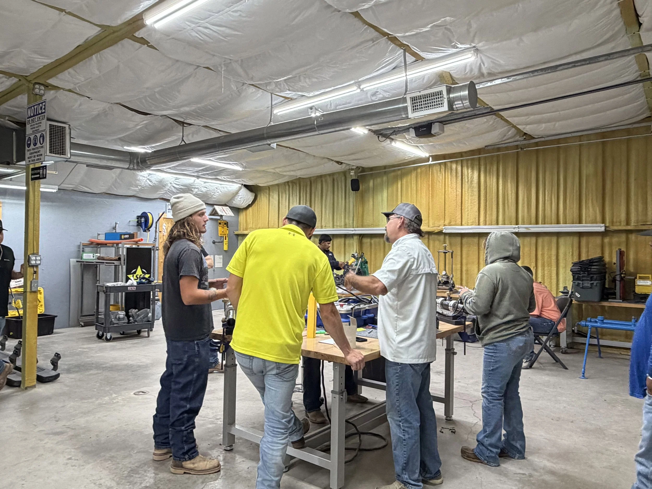 A group of people in casual clothing gathered around a workbench in an industrial workshop. The ceiling is covered with insulation and pipes, and there are various tools and equipment around the space.