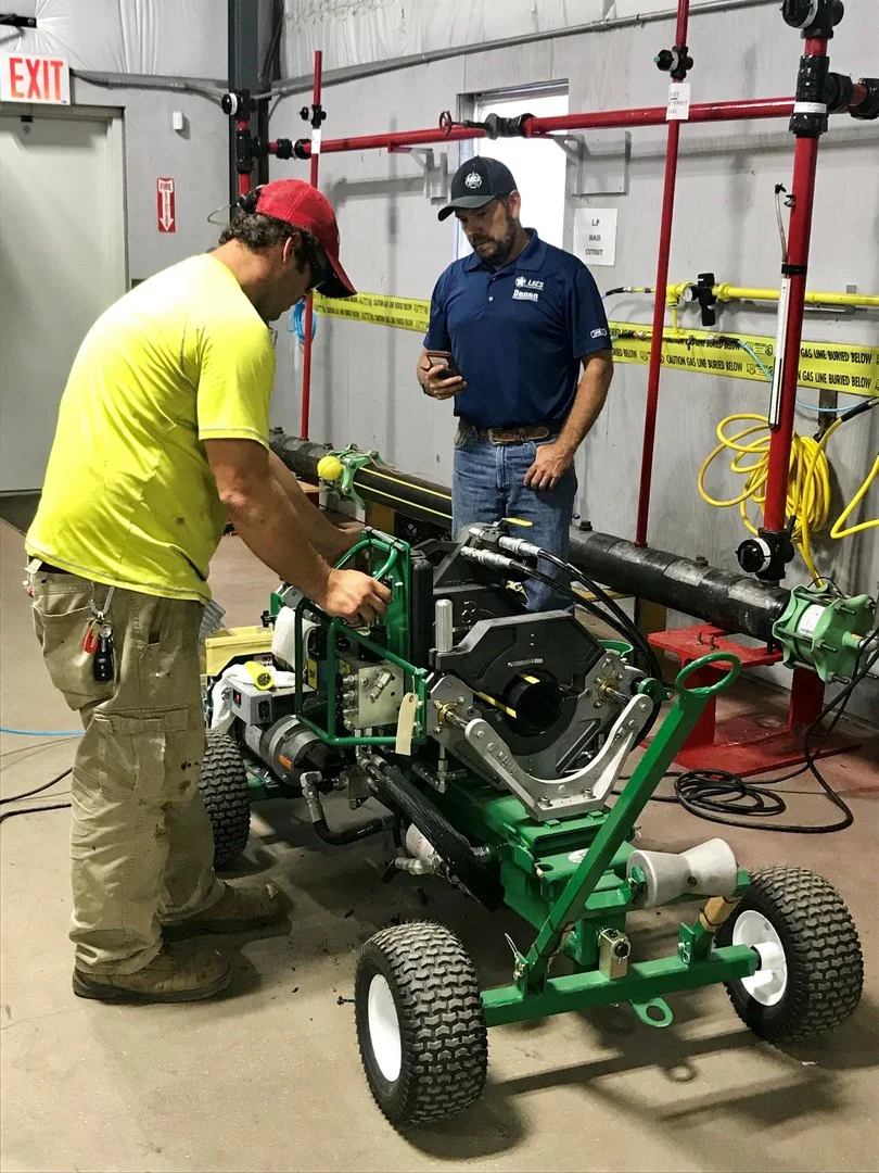 Two men working on a large green equipment with wheels in an industrial setting. One man in yellow shirt and red cap is adjusting the equipment, while another man in blue shirt and dark cap observes.