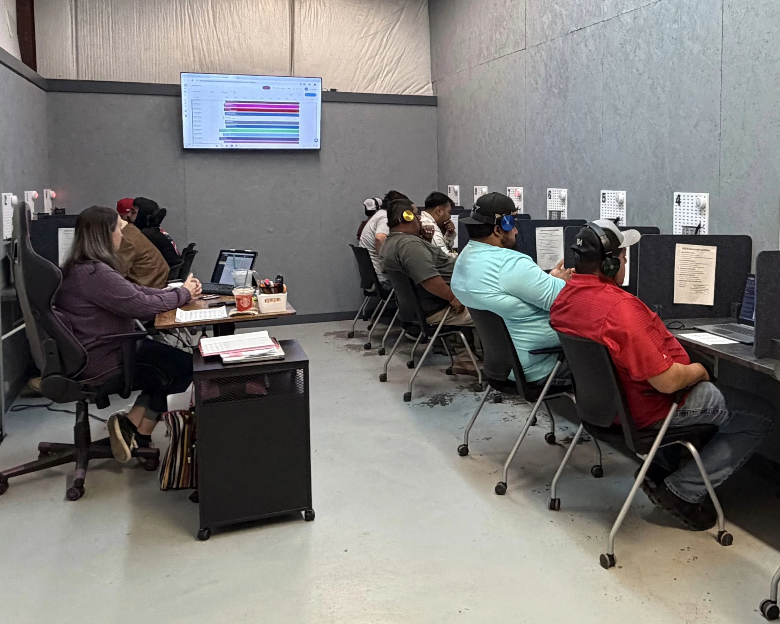 People sitting at desks in a classroom or training room with a large screen displaying a colorful schedule or chart on the wall.