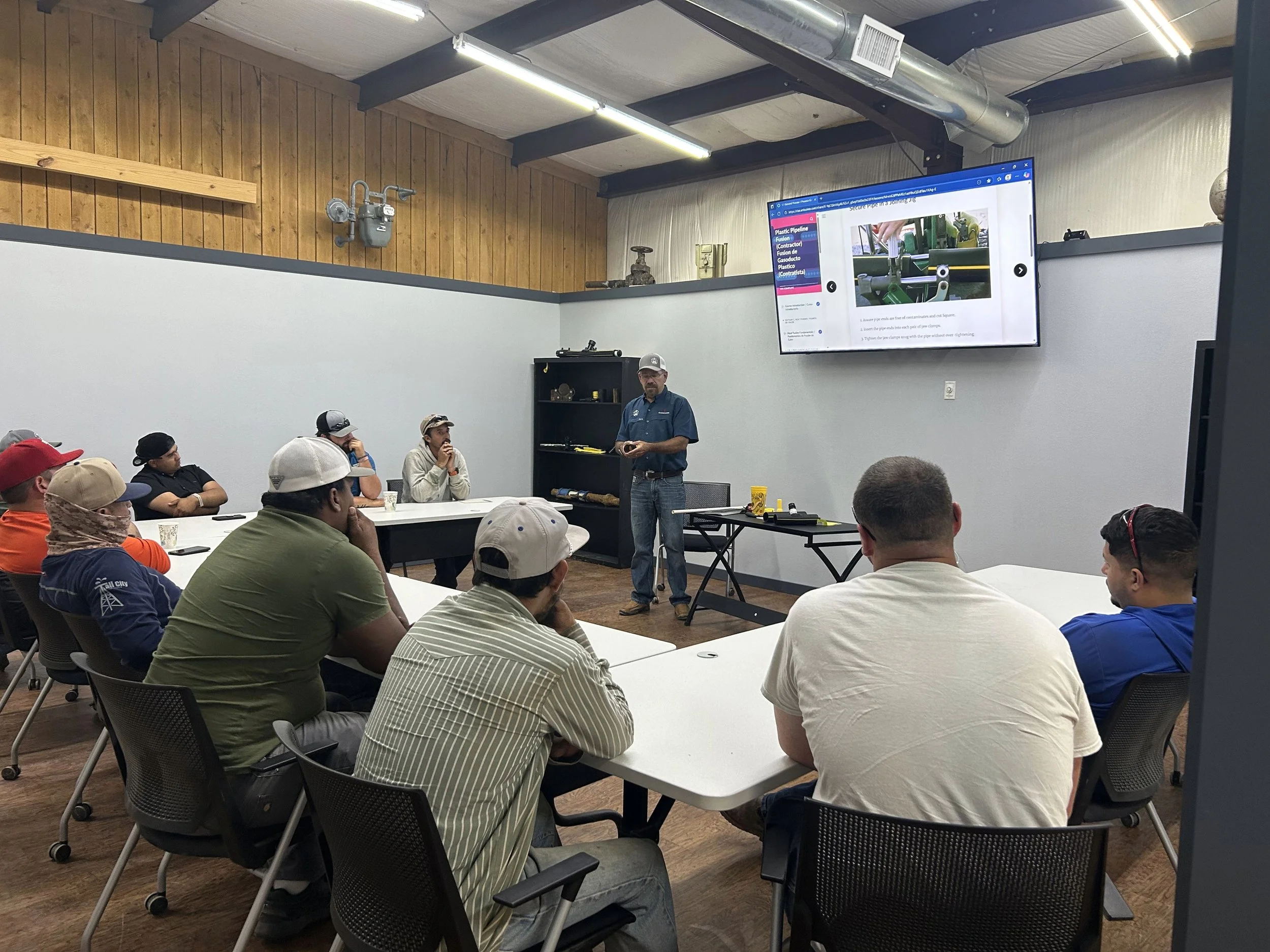 A man in a blue shirt and cap is giving a presentation to a group of people seated at tables in a room with wooden and white walls. A large screen displays a slide related to piping.