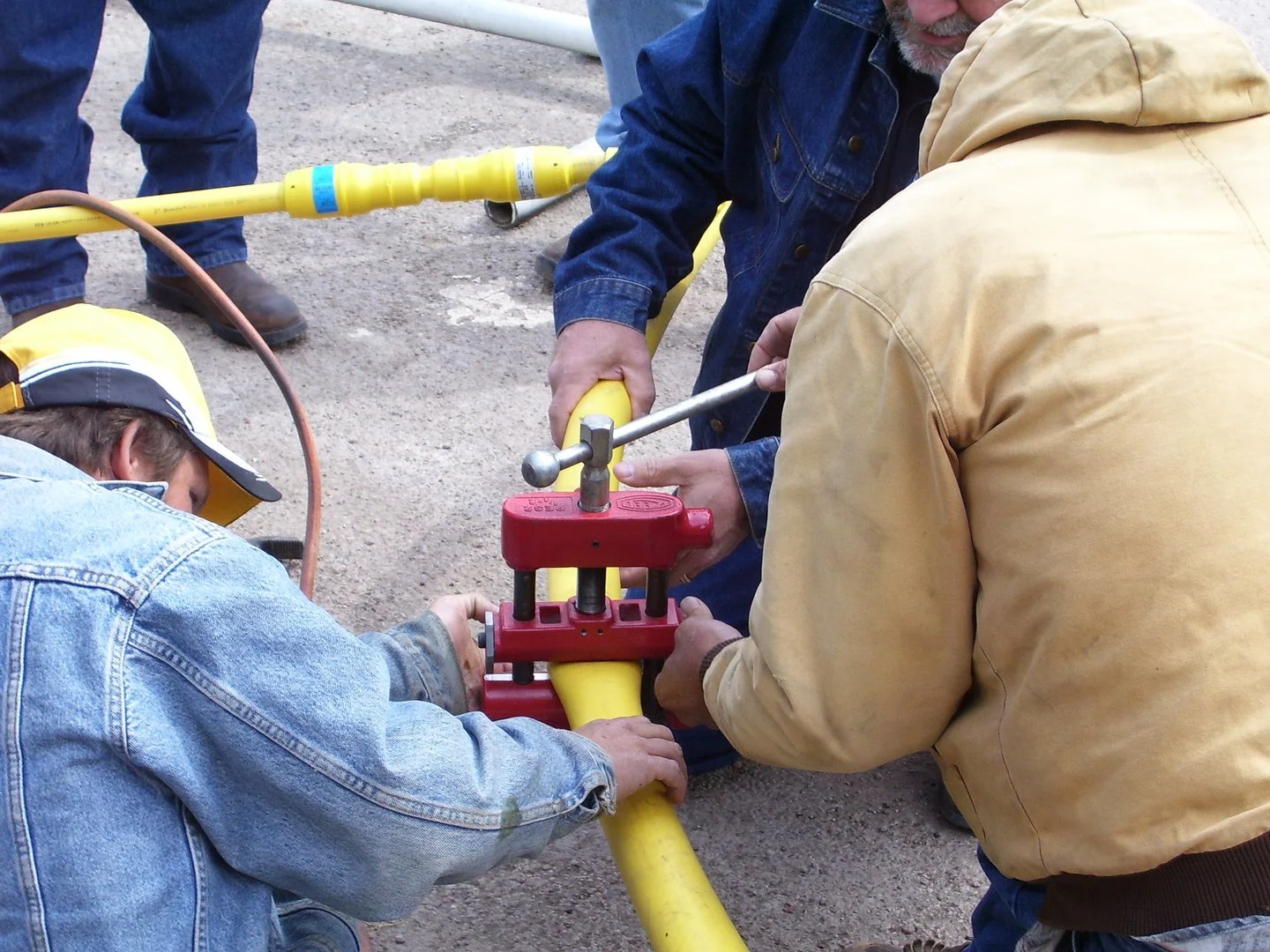 Three people working together to connect yellow pipes in an outdoor setting, with tools and safety equipment.