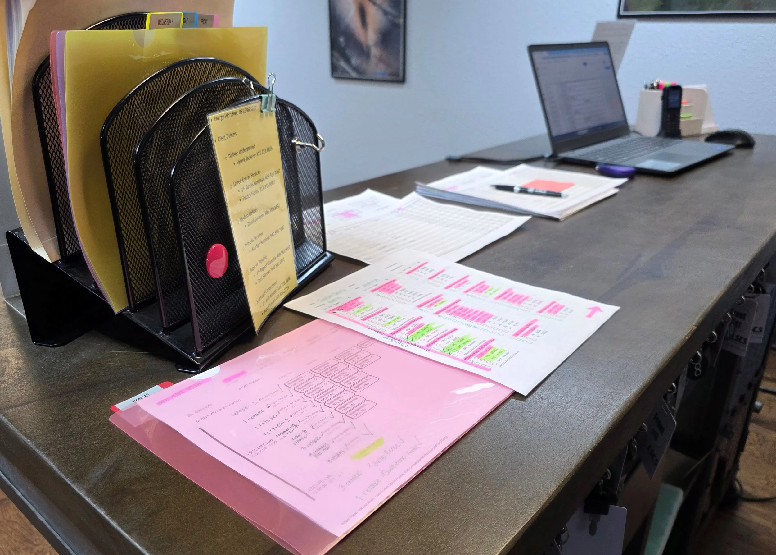 Office desk with pink and yellow folders, printed documents with highlighted sections, a laptop, and office supplies.