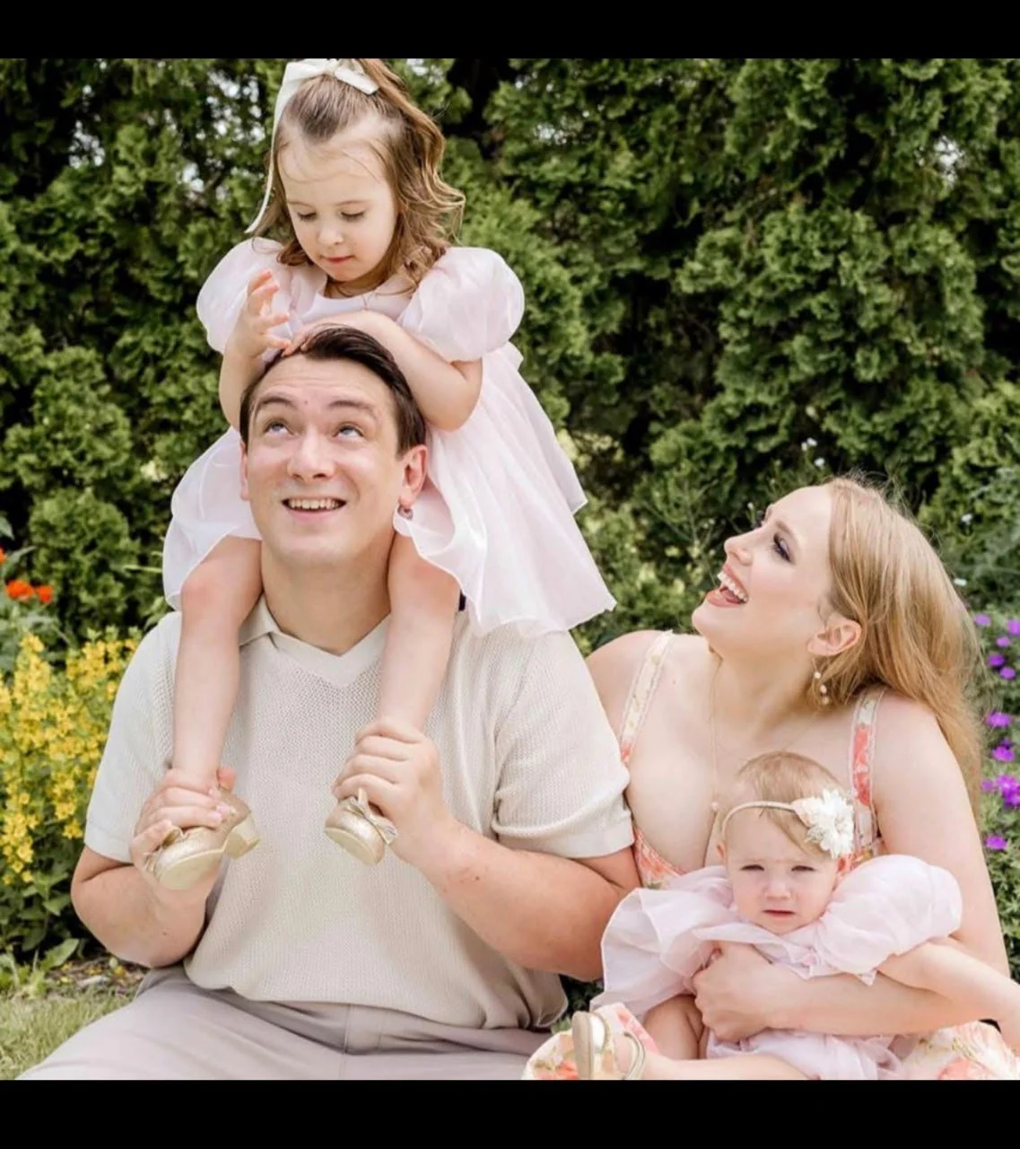 A happy family outdoors on a sunny day, with a man carrying a young girl on his shoulders, a woman smiling at the girl, and a baby girl sitting on the woman's lap, surrounded by greenery and colorful flowers.