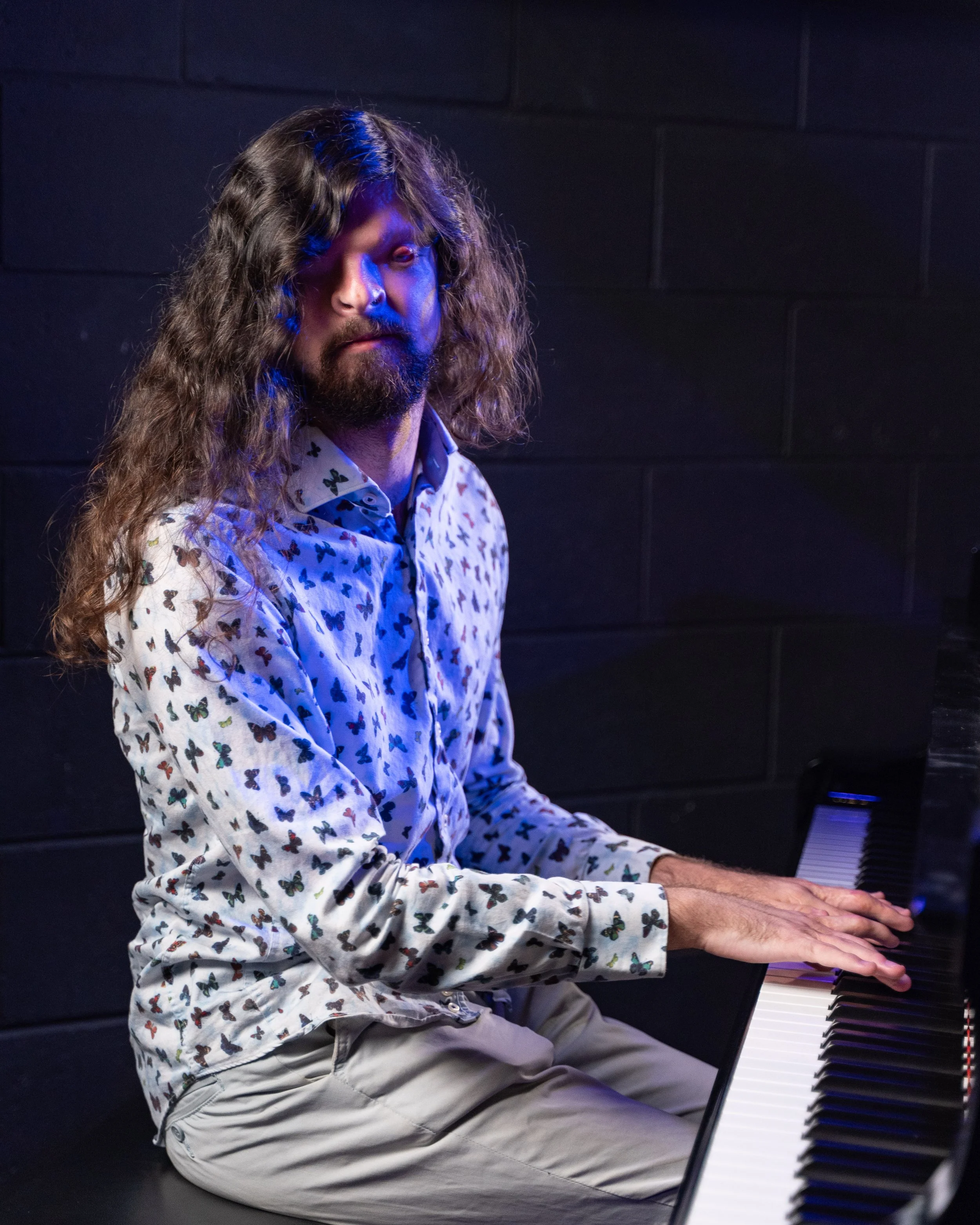 A man wearing a white long-sleeved butterfly patterned shirt with long, curly hair looks at the camera whilst playing a grand piano