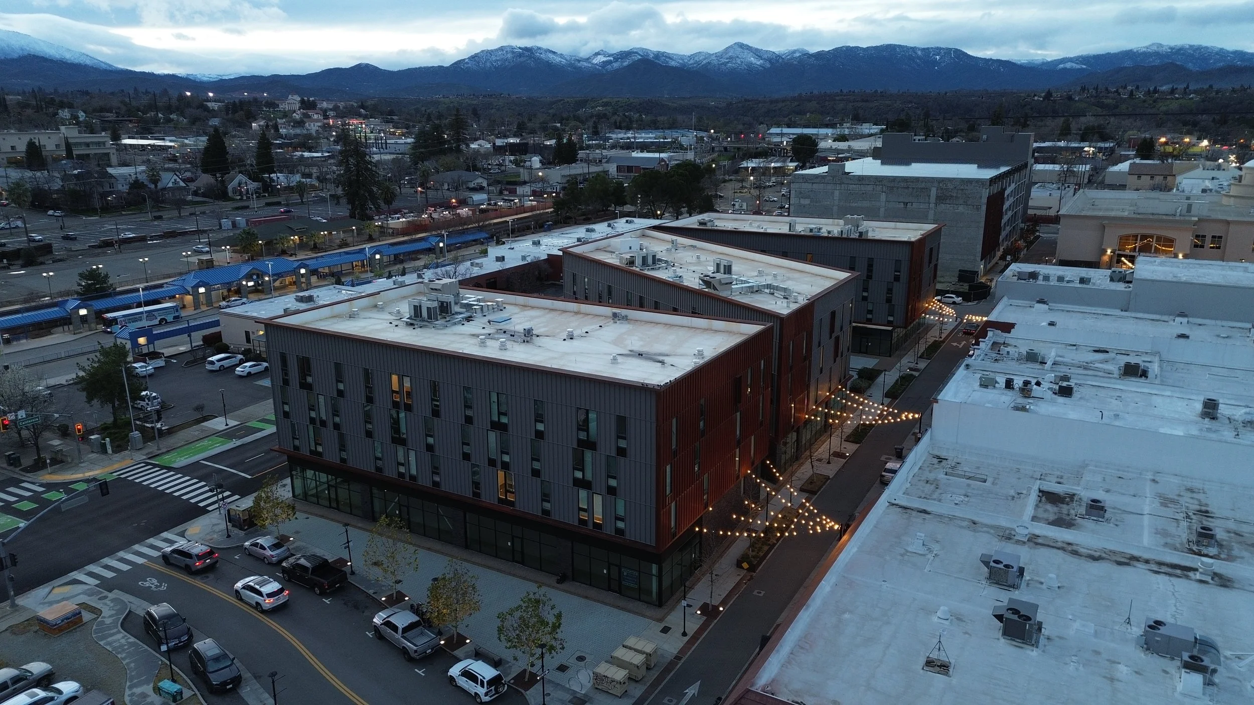 An aerial view of a cityscape at dusk with modern buildings, parked cars, string lights, and mountains in the background.
