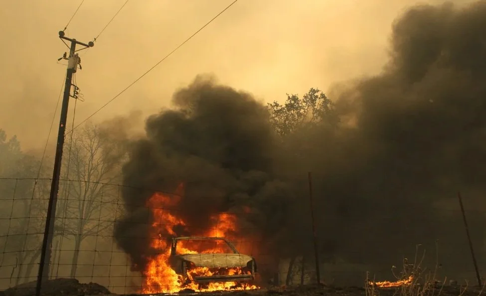 A vehicle is burning with large flames and thick black smoke rising into the sky, near power lines and trees.