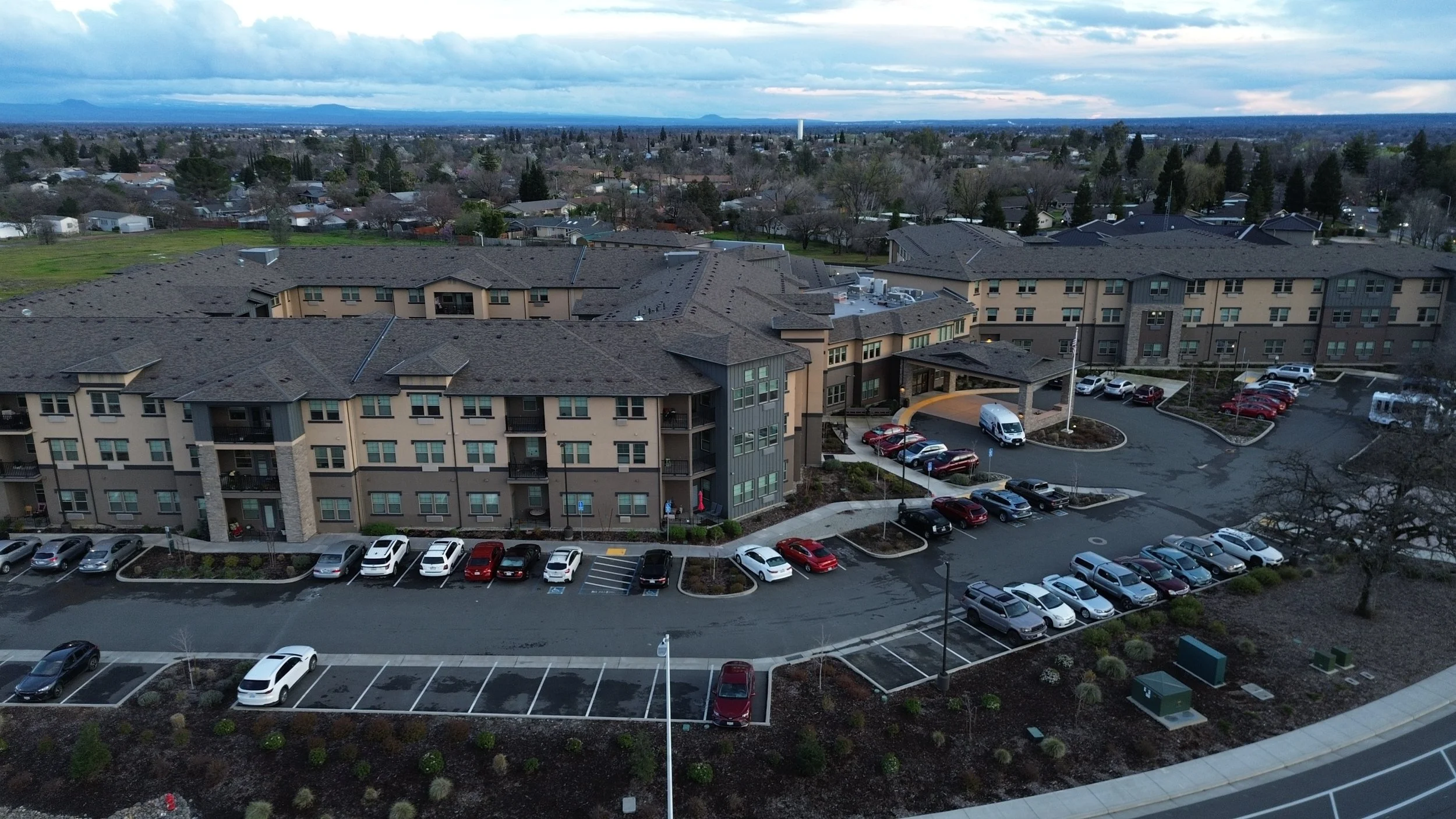 Aerial view of a multi-story apartment complex with parking lots filled with cars, surrounded by trees and a cityscape in the background under a cloudy sky.