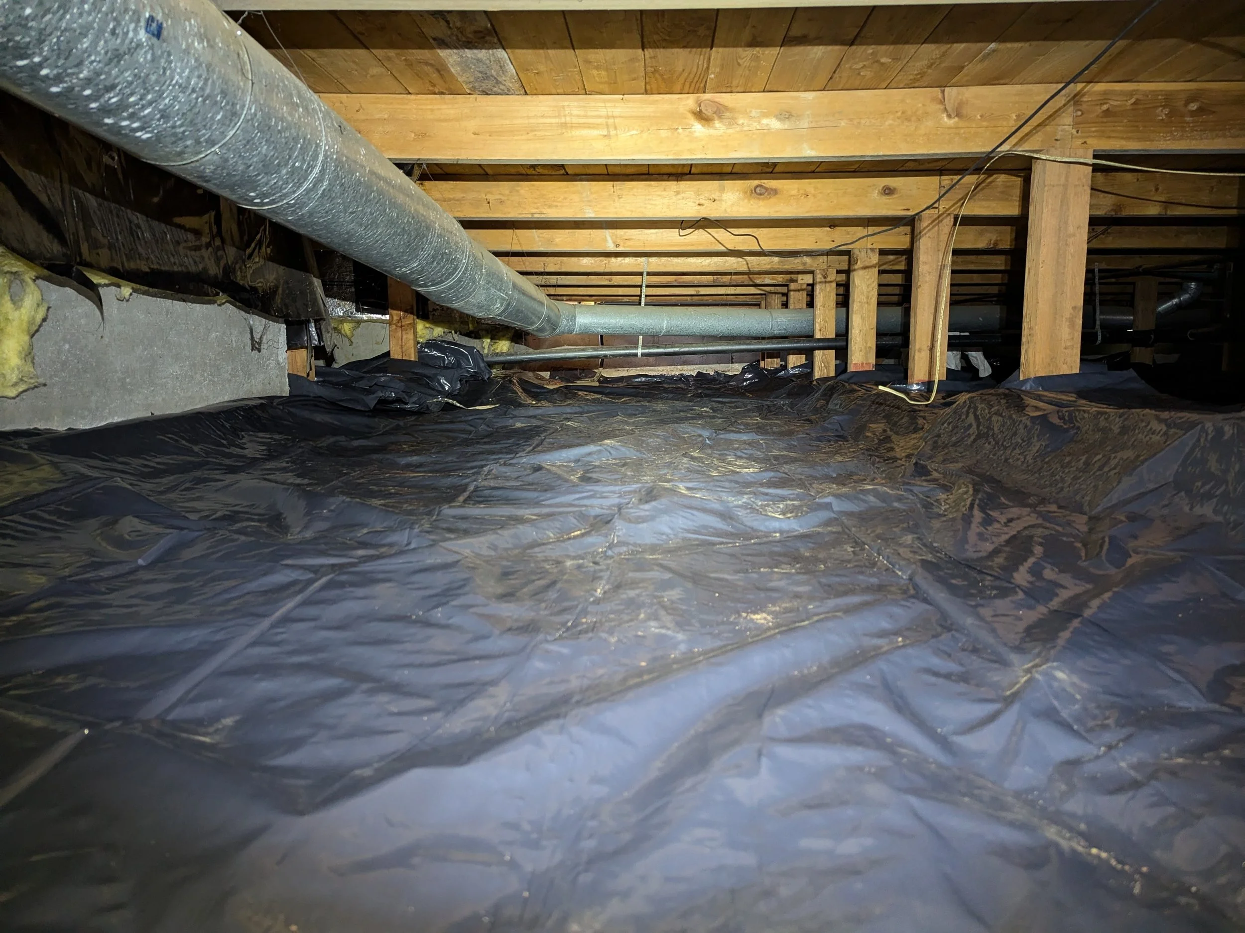 An attic space with exposed wooden beams, a large duct running along the ceiling, and a black plastic vapor barrier on the floor.