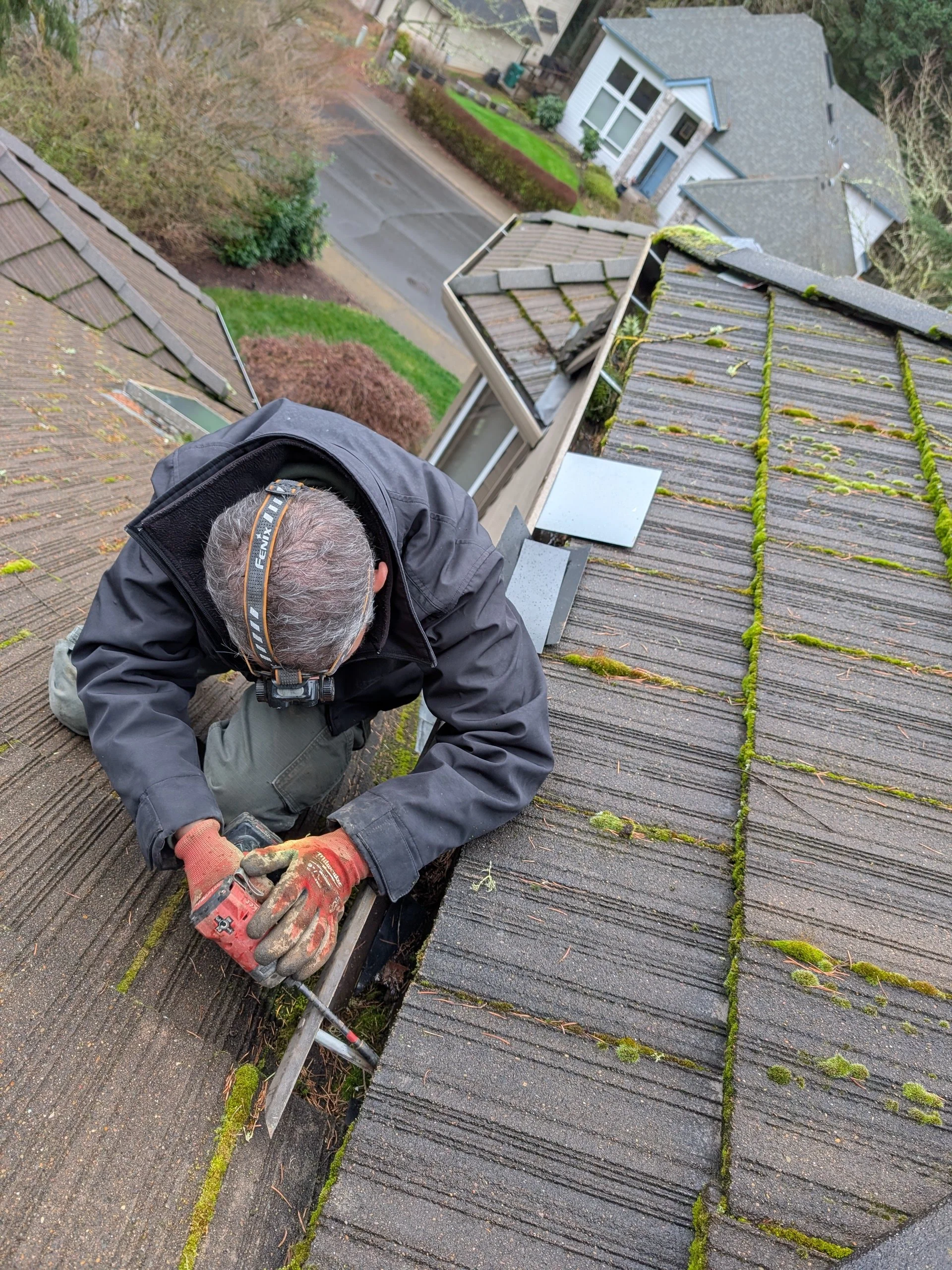 A man working on a roof, using a tool to repair or inspect the gutter, with residential houses and a street visible in the background.