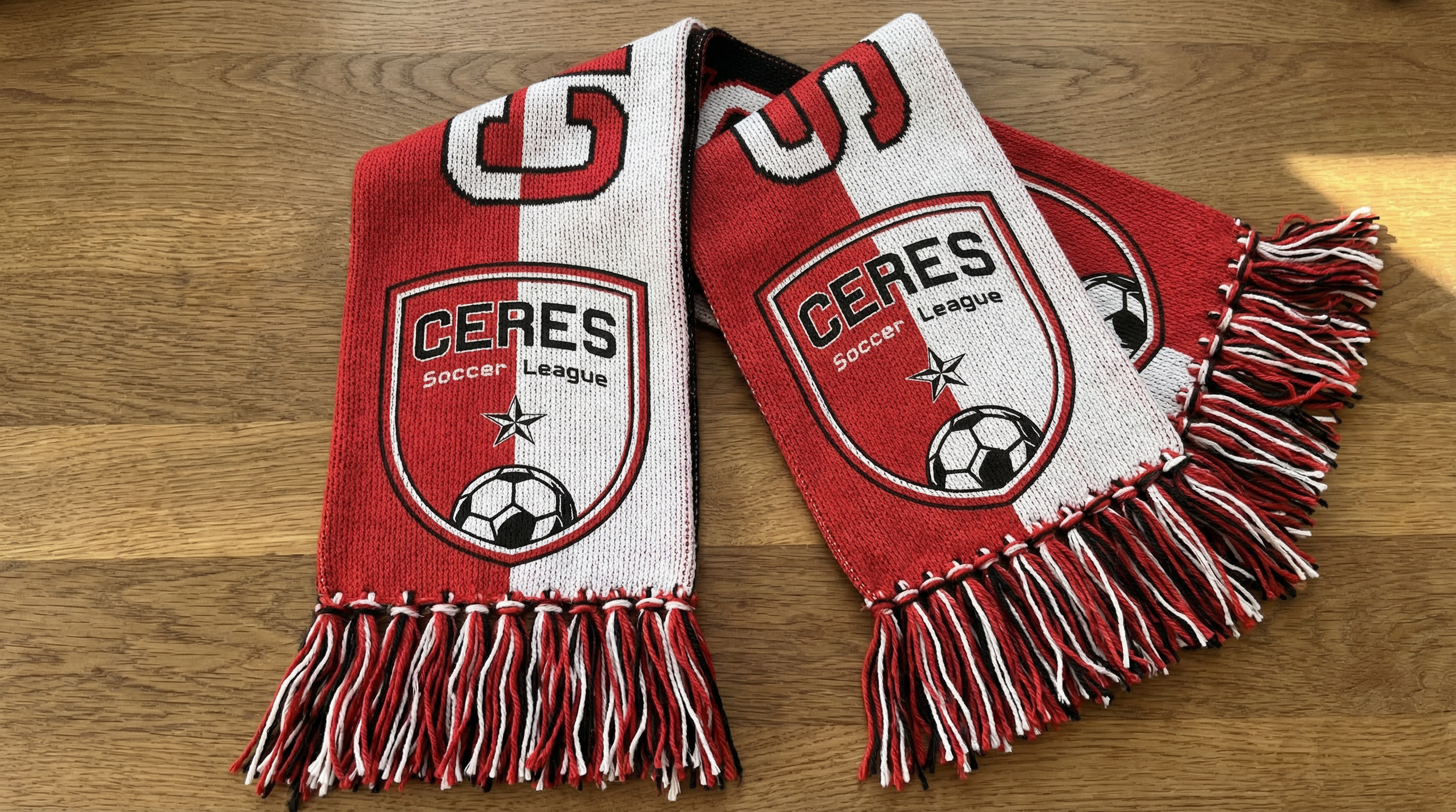 Red and white Ceres Soccer League scarf with fringe, featuring a soccer ball logo and the league name on a wooden surface.