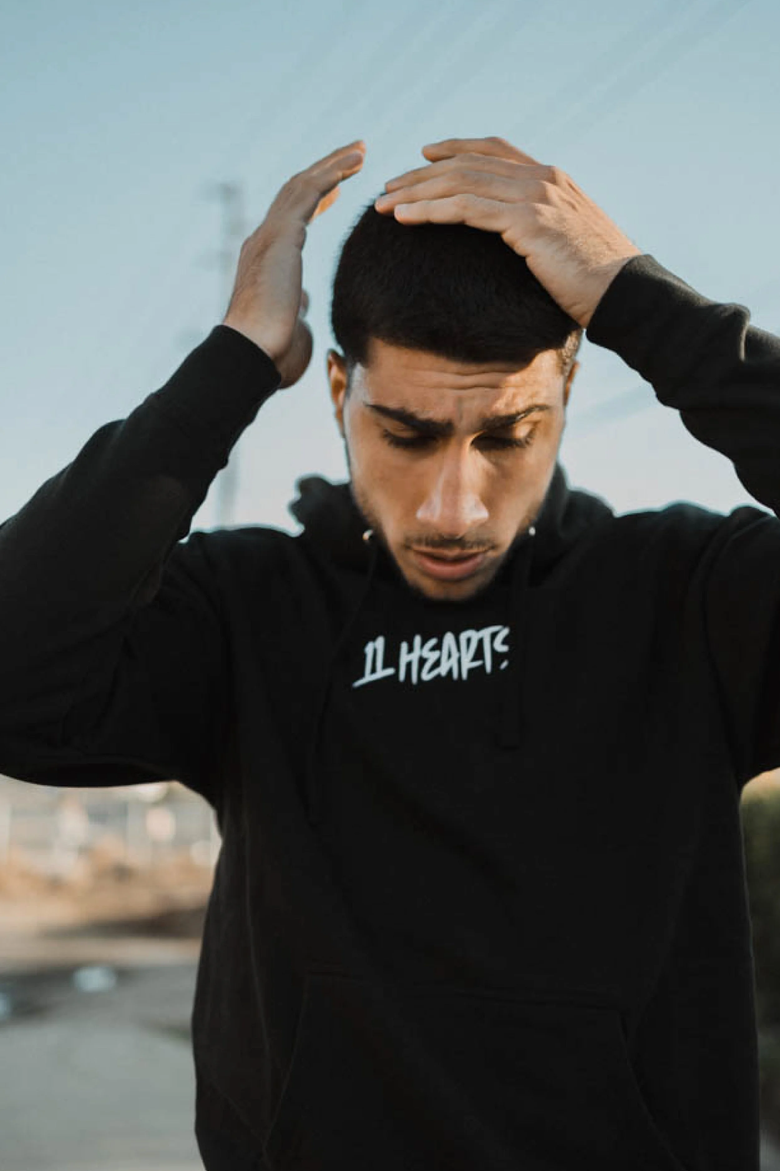 A young man with dark hair and light skin is holding his head with both hands, wearing a black hoodie with white text "11 Hearts".