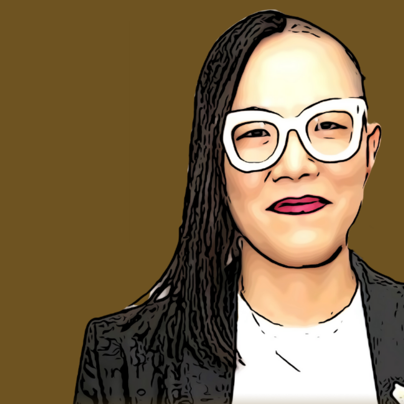 Woman with long dark hair, white glasses, red lipstick, wearing a black blazer and white top, against a brown background.