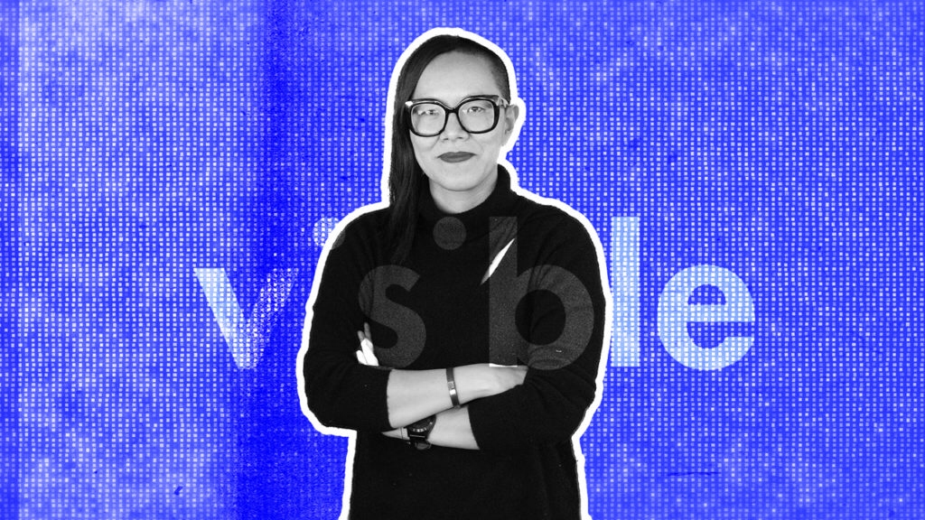 A woman with shoulder-length dark hair, wearing glasses, a black turtleneck, and a watch, standing with arms crossed in front of a digital blue background with the barcode and the word 'visible' in faded white letters.