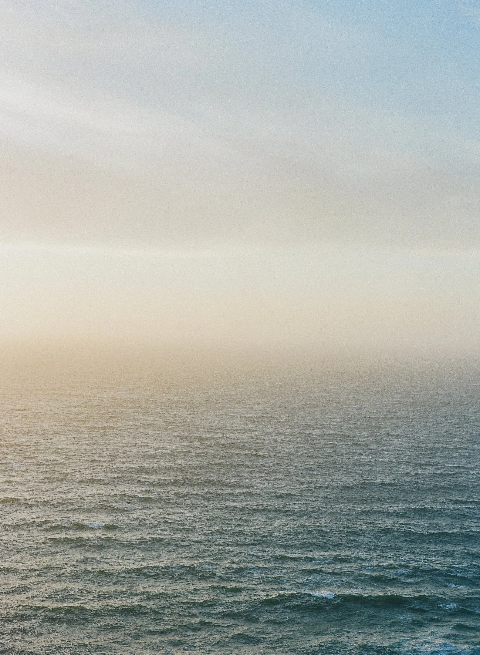 Calm ocean waters with gentle waves under a horizon with sky and clouds in muted colors.