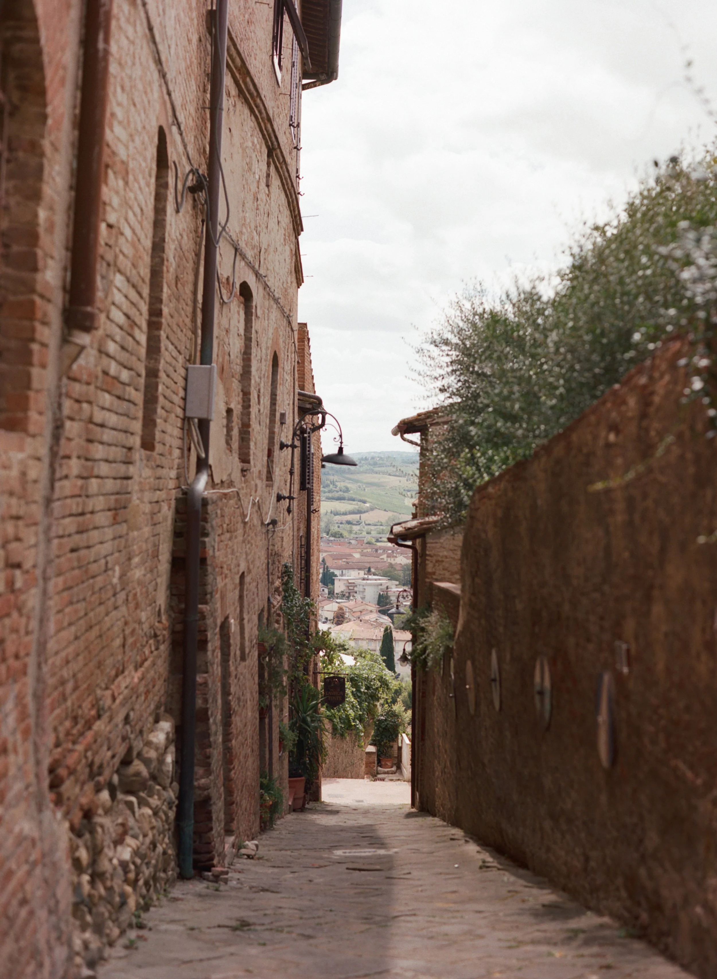 A narrow cobblestone alleyway between old brick buildings with lush greenery and a view of a town and rolling hills in the distance, under a cloudy sky.