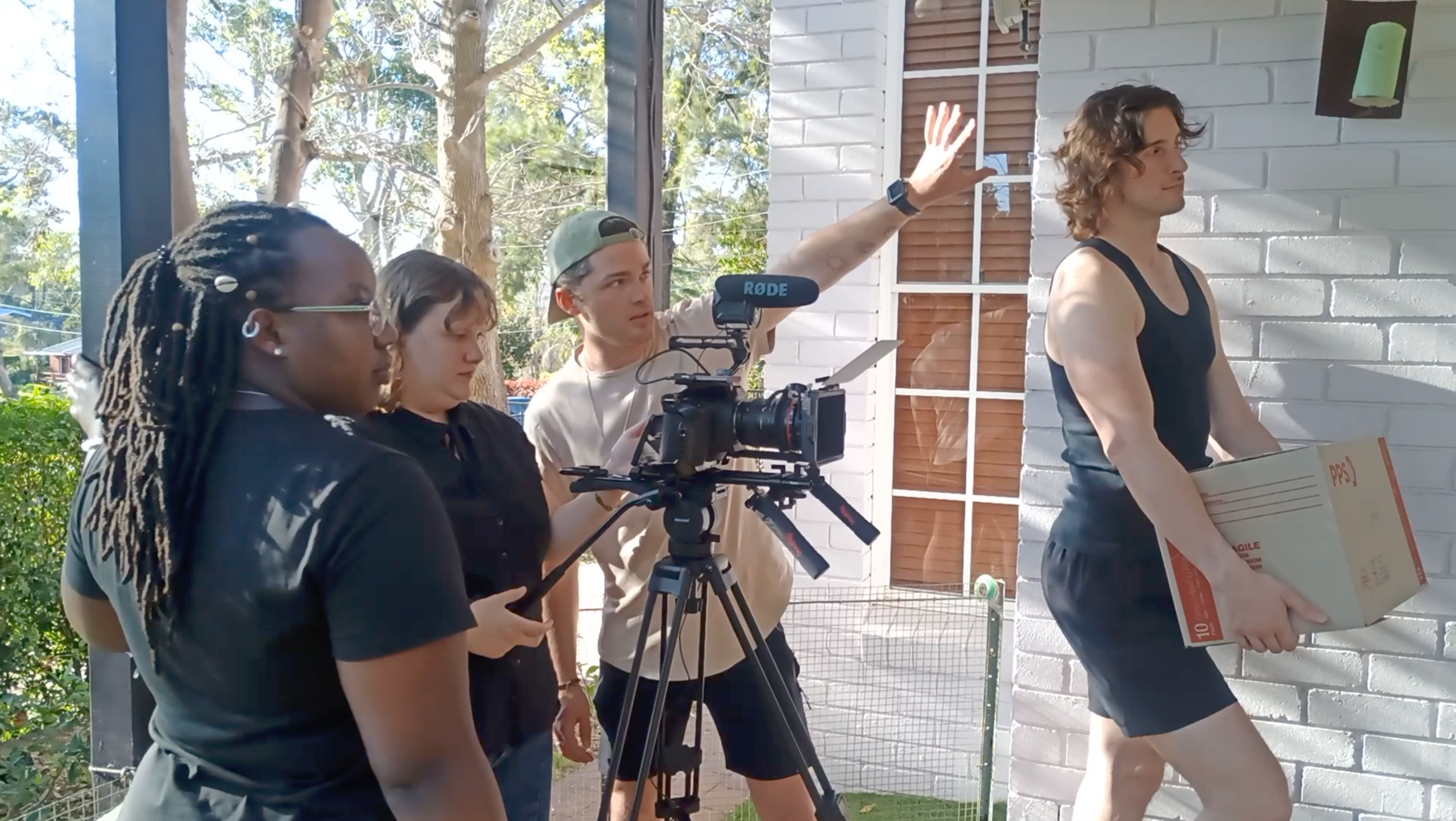 A film crew filming a man wearing a black tank top and shorts carrying a cardboard box outside near a white brick wall.