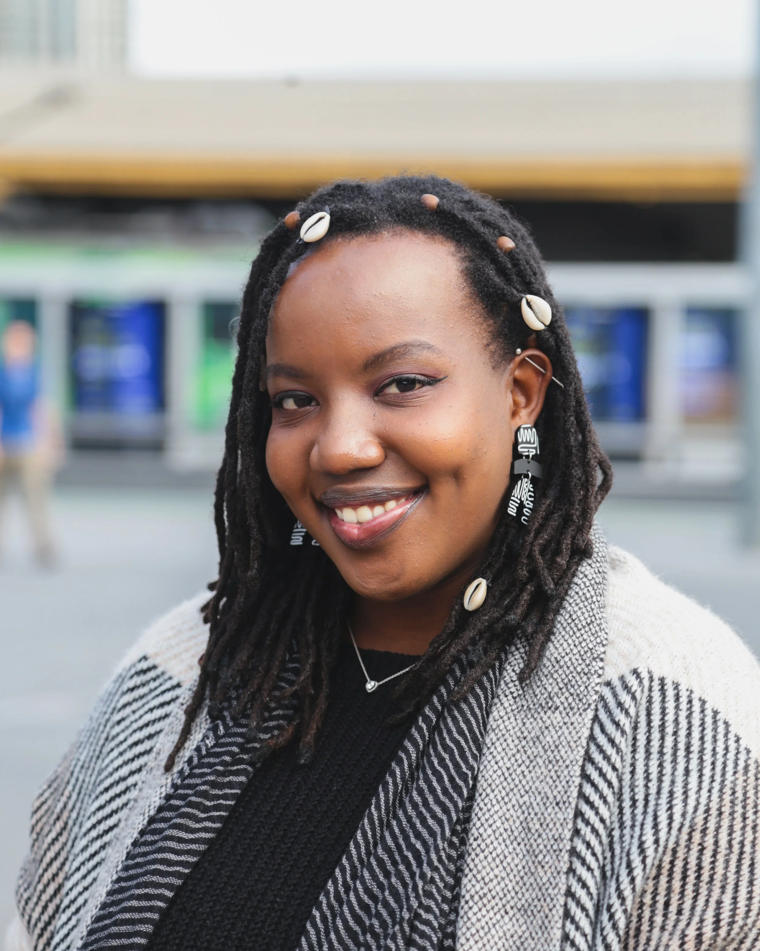 A smiling woman with dreadlocks decorated with seashells, wearing earrings and a black top with a patterned jacket, standing outdoors in an urban area.