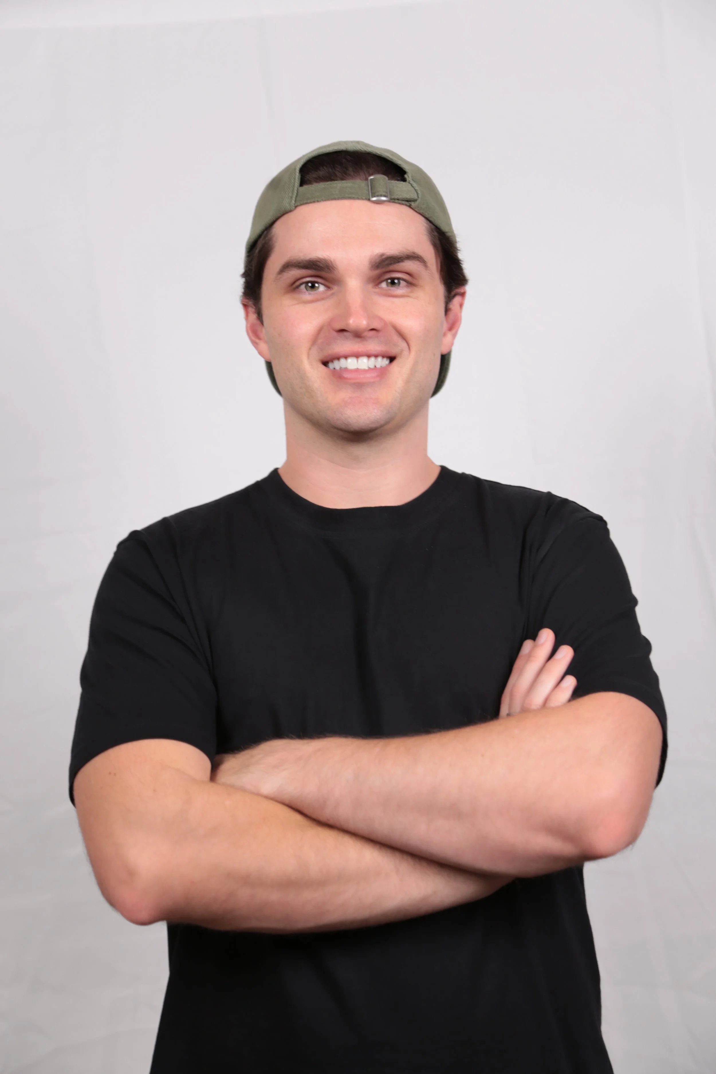 A young man with dark hair and a broad smile, wearing a black t-shirt and a green baseball cap backwards, standing with arms crossed in front of a plain white background.