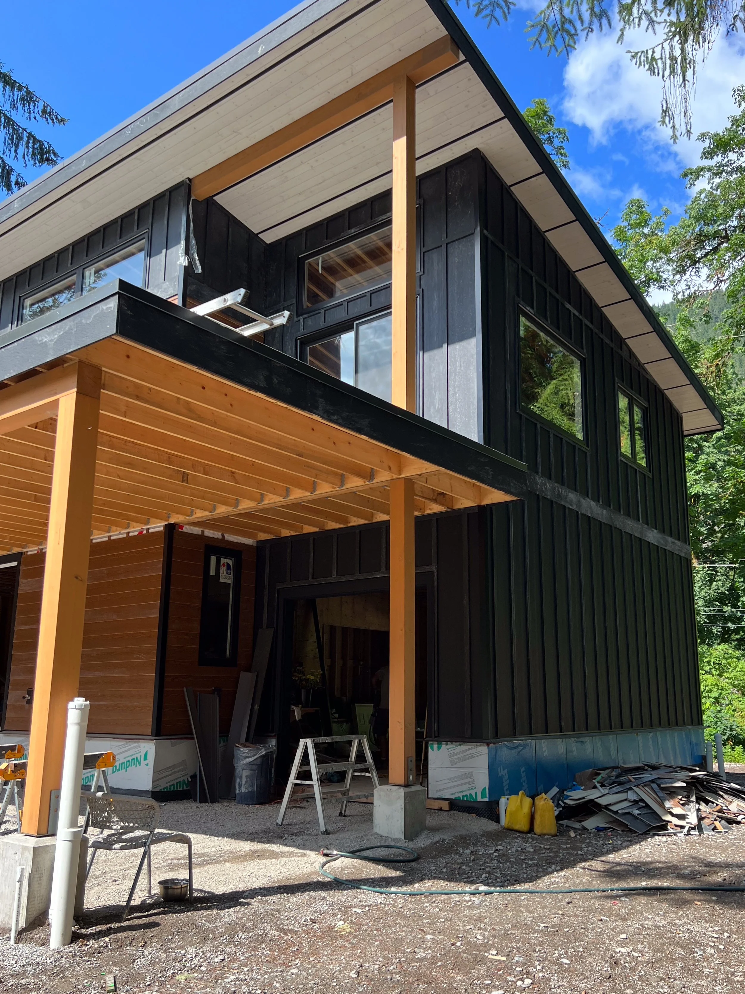 Under construction modern two-story house with black and natural wood exterior, large windows, and a partially built porch with wooden beams and construction materials around.