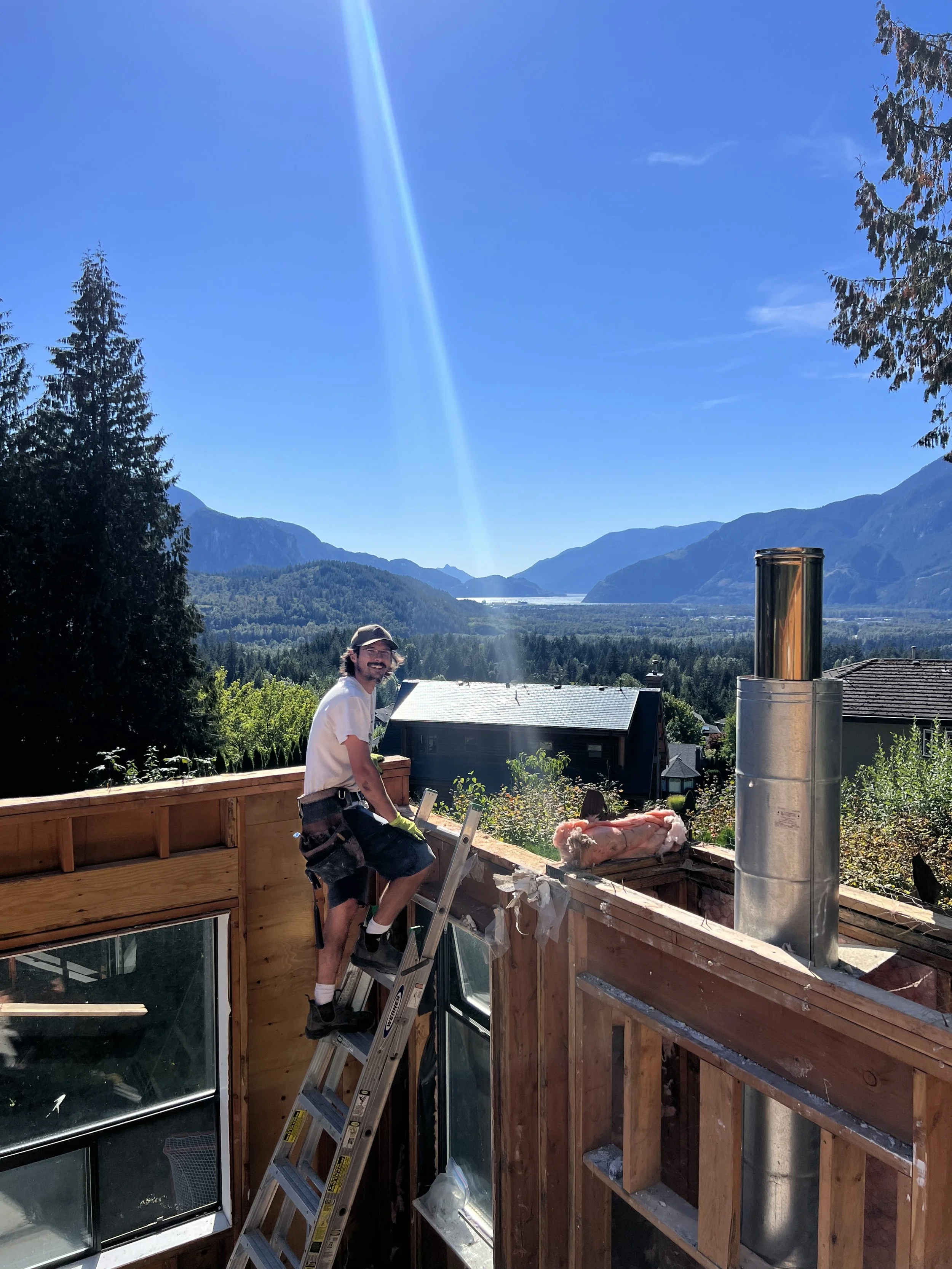 A man standing on a ladder on a construction site, working on a wooden frame of a building, with mountains and trees in the background under a clear blue sky.