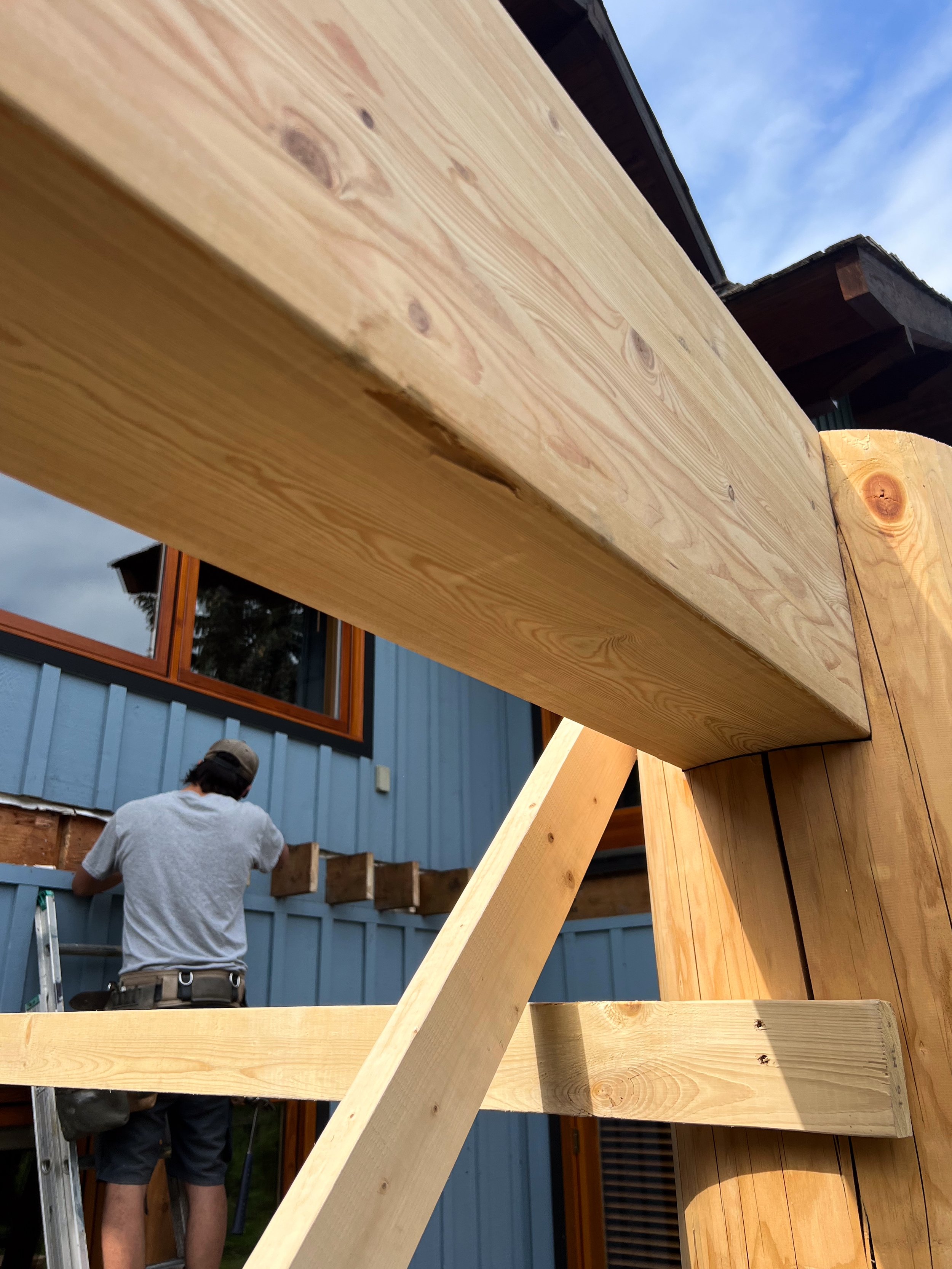 Close-up of a wooden beam under construction with a person working in the background on a house with blue siding and orange window frames.