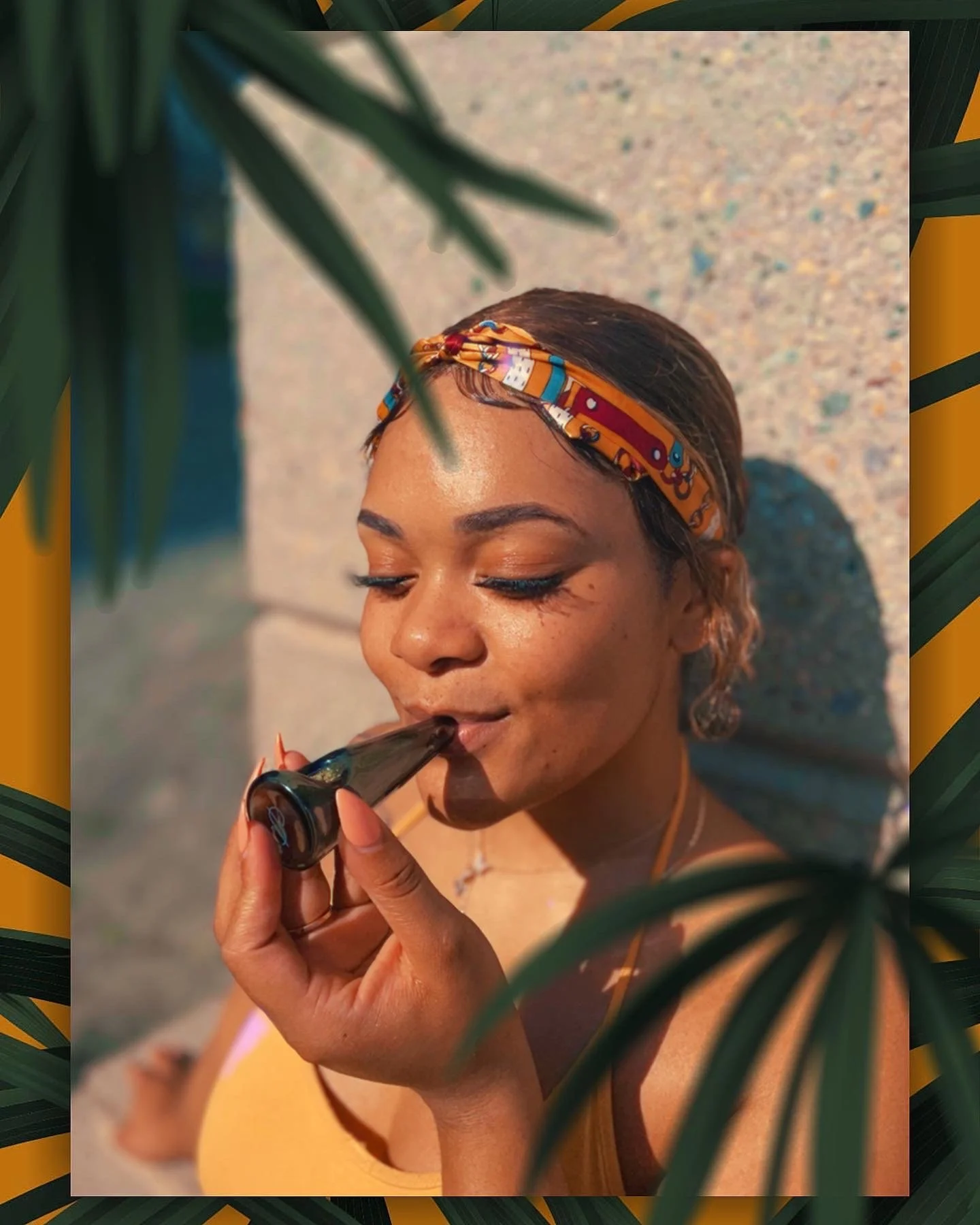 Young woman with a colorful headband enjoying a clear pipe outdoors near a sandy surface, with green leaves partially obscuring the view.