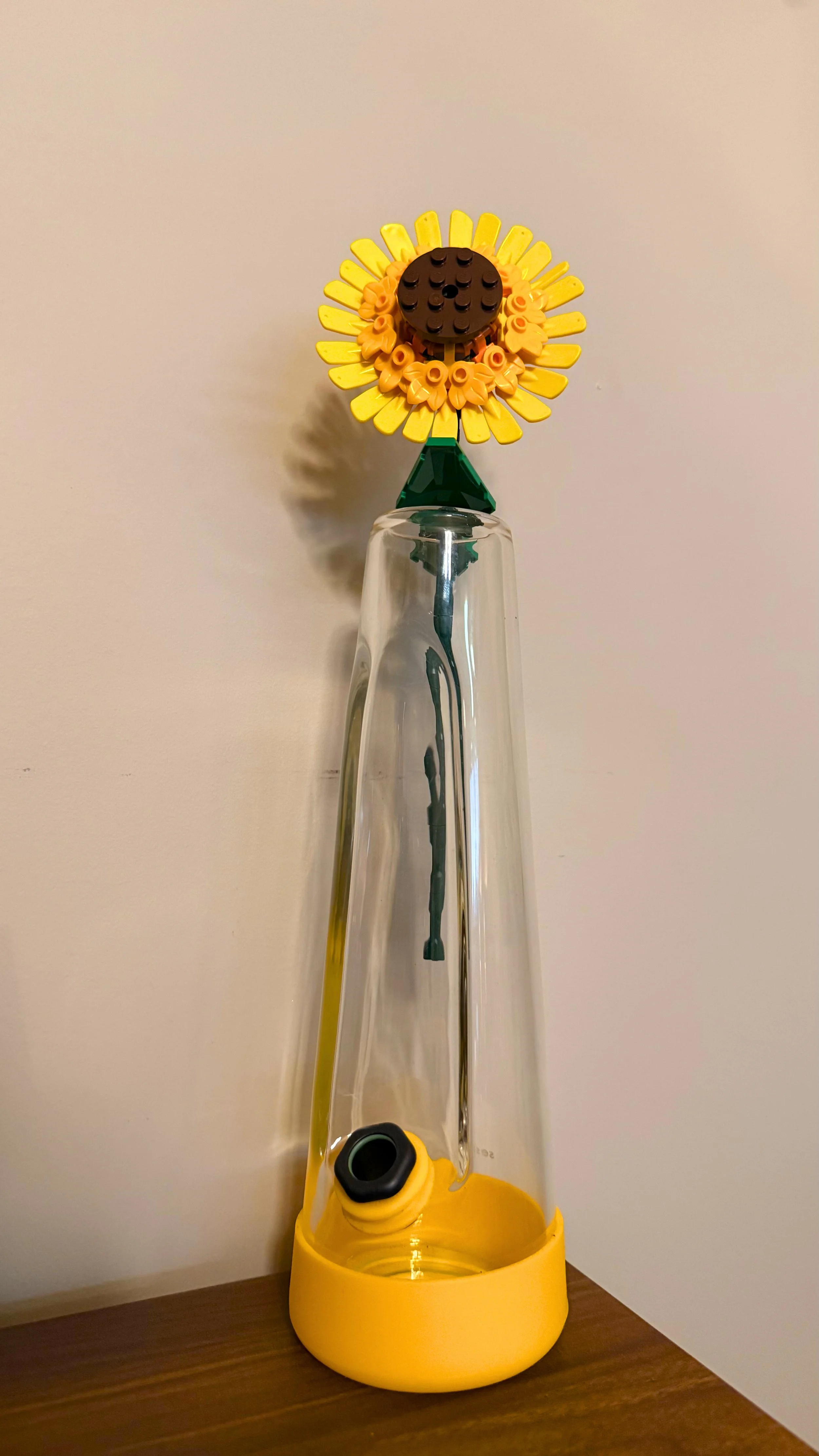 A decorative flower made from LEGO pieces with yellow petals, a brown center, and an orange ring of smaller pieces, mounted on a glass bong with a yellow base. The flower has a green LEGO stem and leaves.