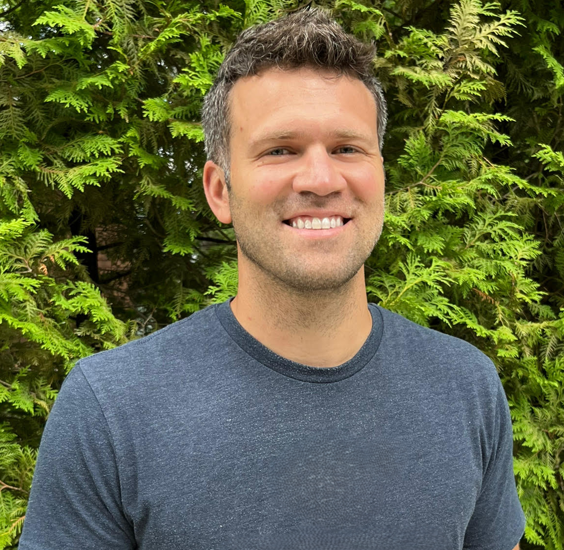 A smiling man with short dark hair and a stubble beard wearing a dark gray t-shirt standing outdoors in front of lush green foliage.