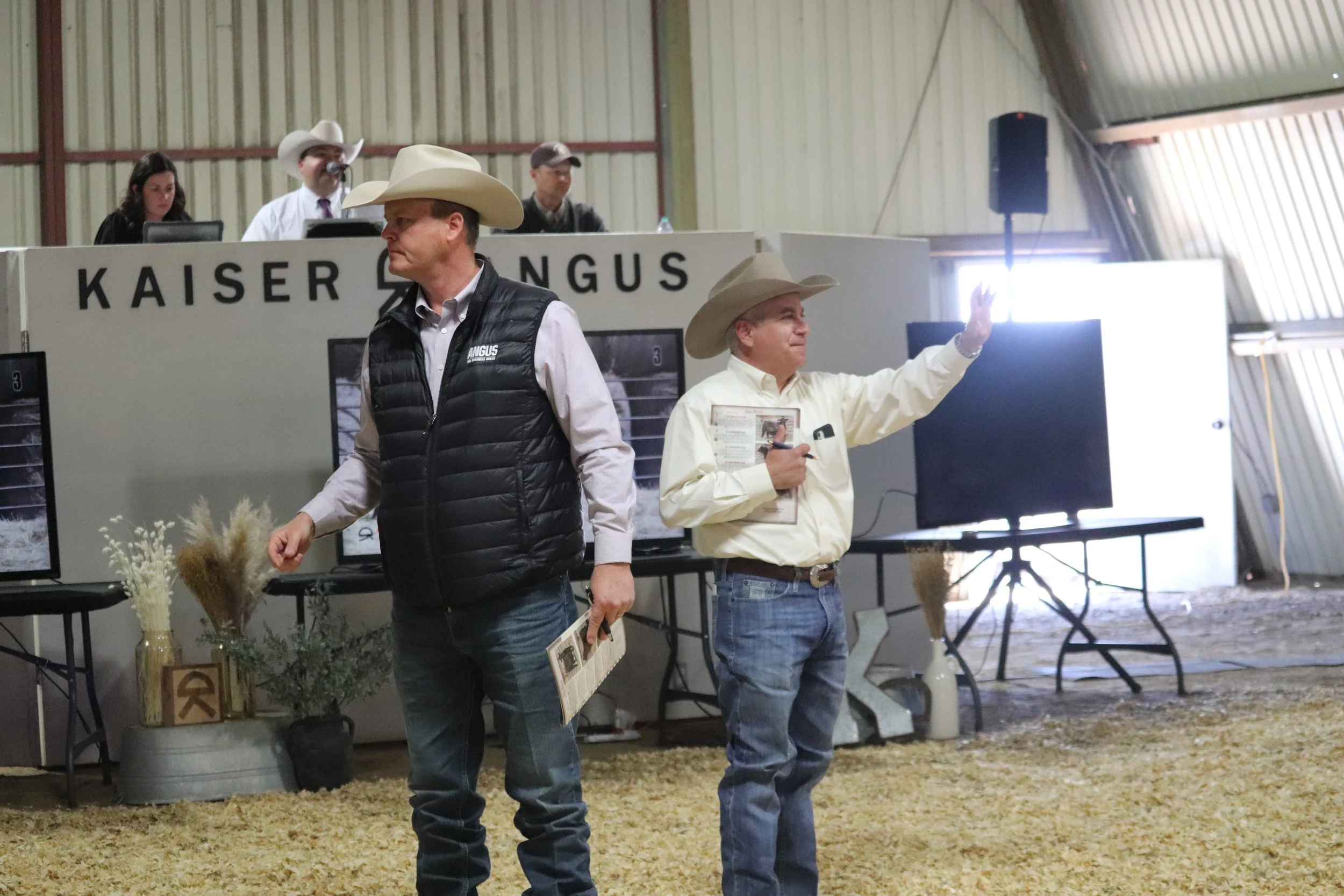 Two men in cowboy hats and dressed in western attire standing in an indoor cattle auction. One man is holding papers and the other appears to be giving a wave. Behind them, a panel of people and multiple screens are visible.