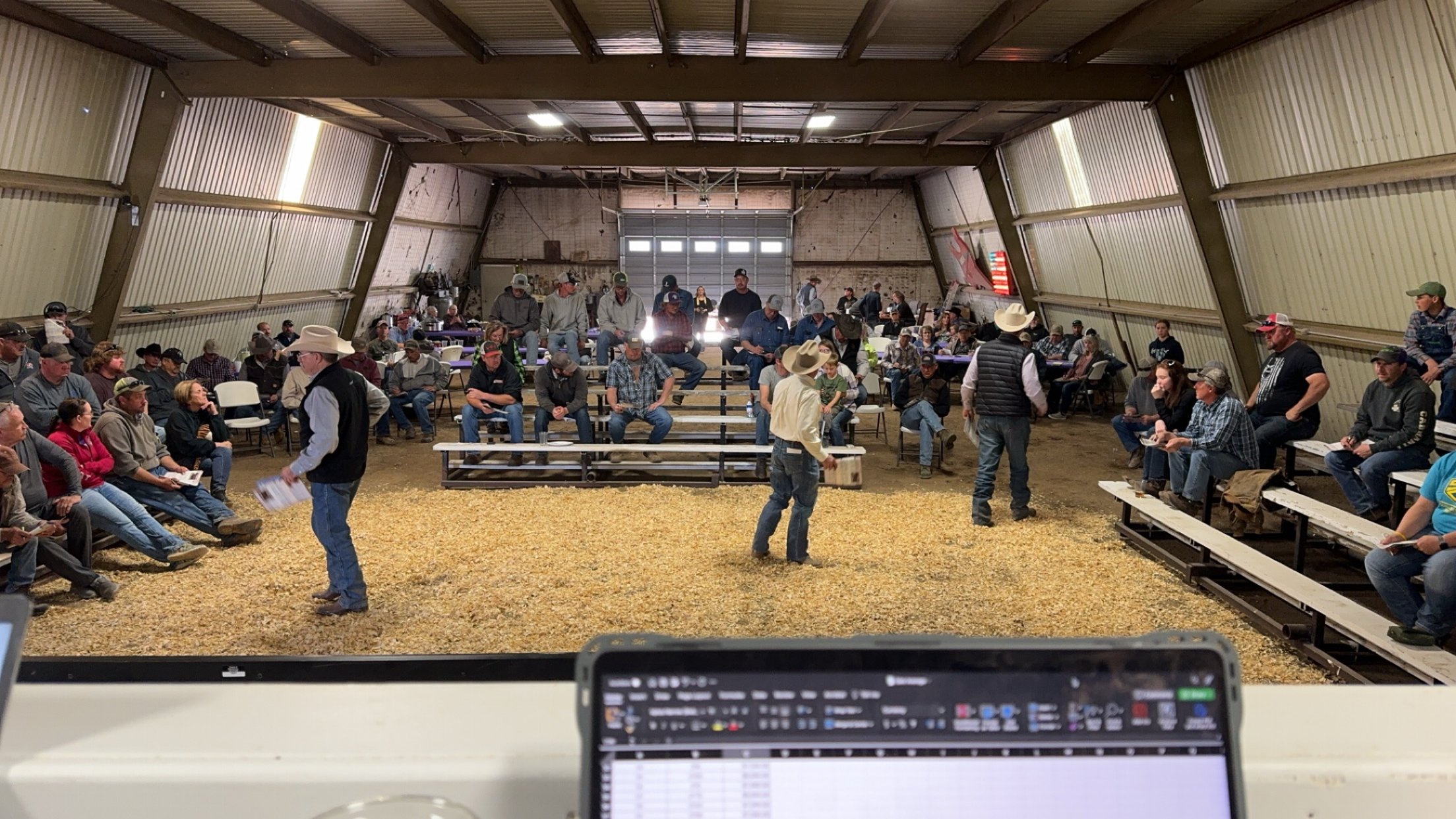 People gathered inside a large barn for a cattle auction, sitting on bleachers and chairs, with some people standing and walking around.