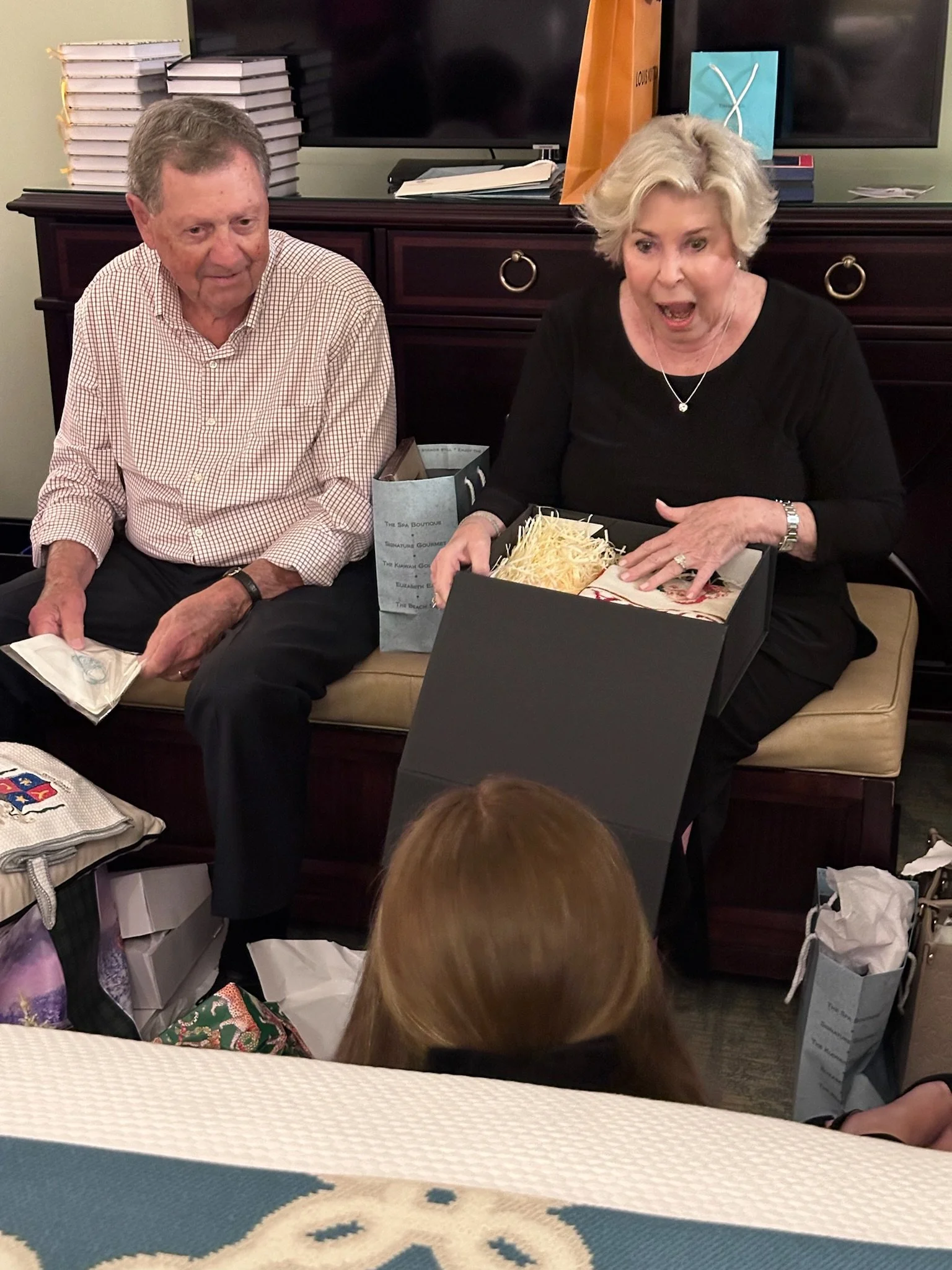 An older man and woman sitting on a bench, exchanging gifts during a celebration. The woman has her mouth open in surprise as she holds an open gift box.