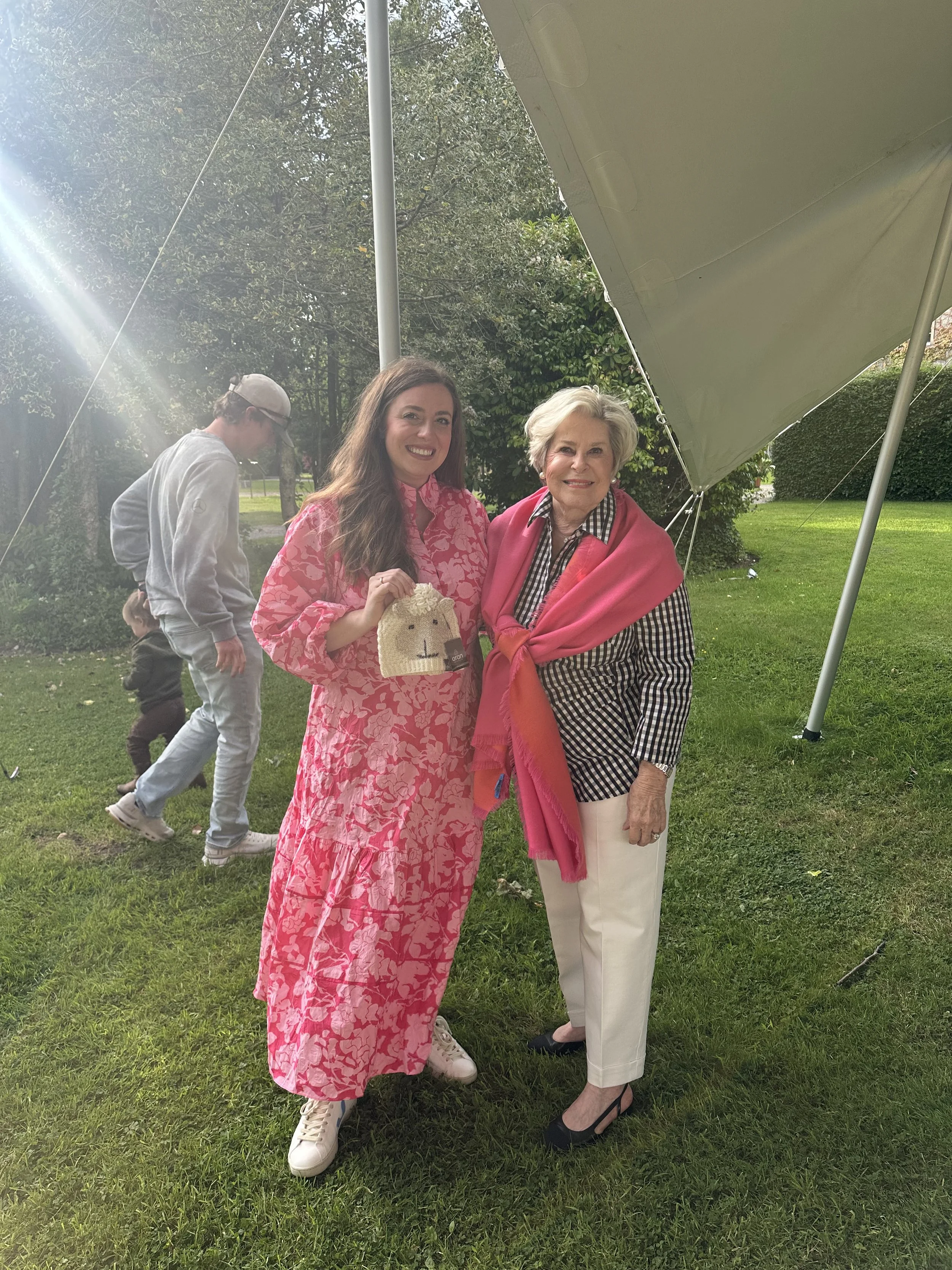 Two women smiling outdoors under a canopy, with a man and child in the background walking on the grass. One woman is wearing a pink floral dress and holding a small beige bag, while the other is in a checkered shirt with a pink shawl.