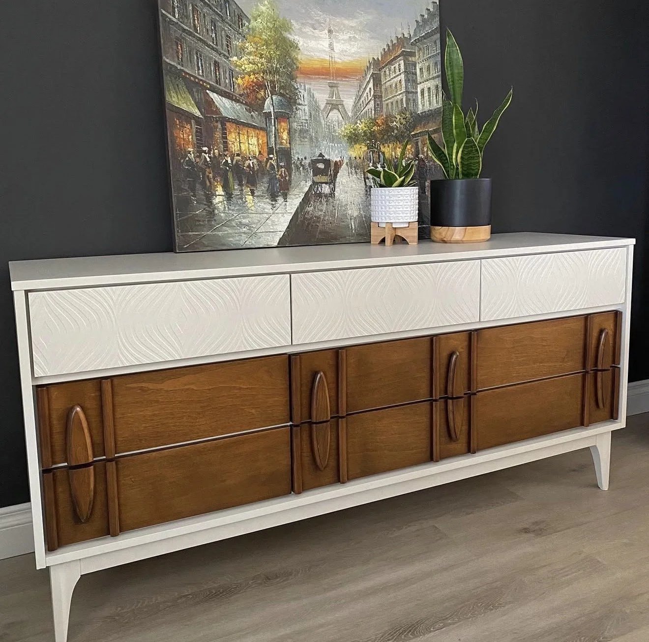 A mid-century modern sideboard with a white top and wood brown cabinet doors, placed against a black wall. On top, there are two potted plants and a framed painting of a rainy cityscape with the Eiffel Tower in the background.
