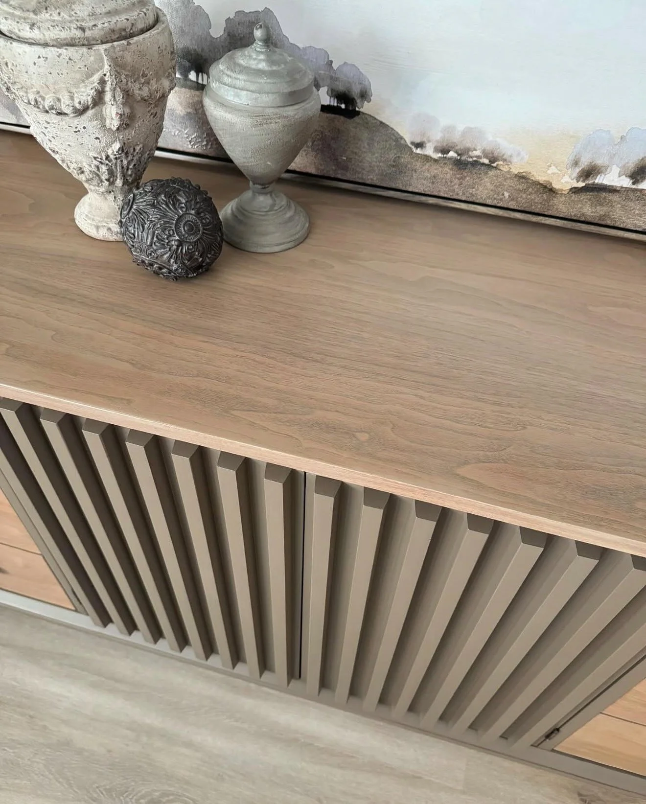Decorative vases and a textured sphere on a wooden cabinet with slatted doors, against a wall with a landscape painting.